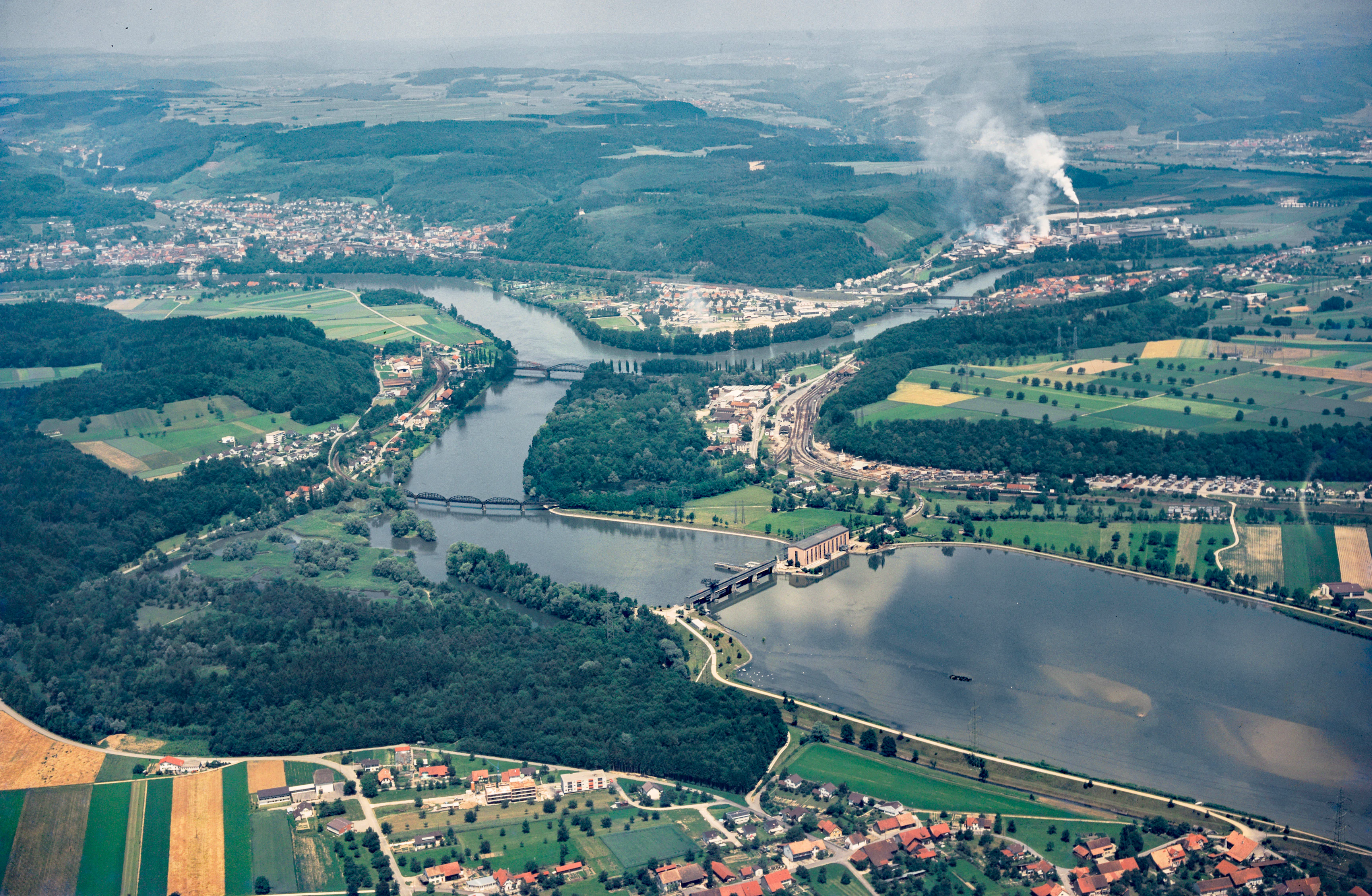 Una vista aérea que muestra un amplio paisaje fluvial en Suiza con la central eléctrica de Klingnau, riberas boscosas, campos agrícolas y pequeños asentamientos residenciales.