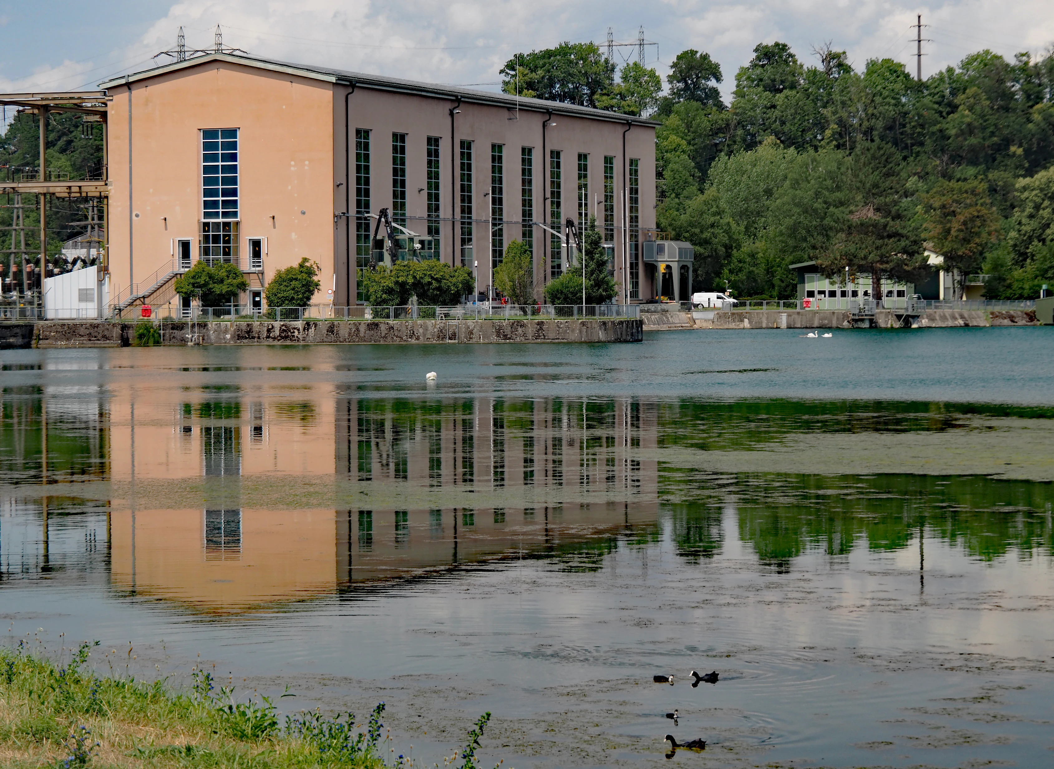 Un gran edificio industrial rectangular con paredes de color melocotón claro y múltiples ventanas verticales se alza en la orilla de un lago tranquilo, con su imagen reflejada en el agua, y árboles y una torre de tendido eléctrico al fondo.