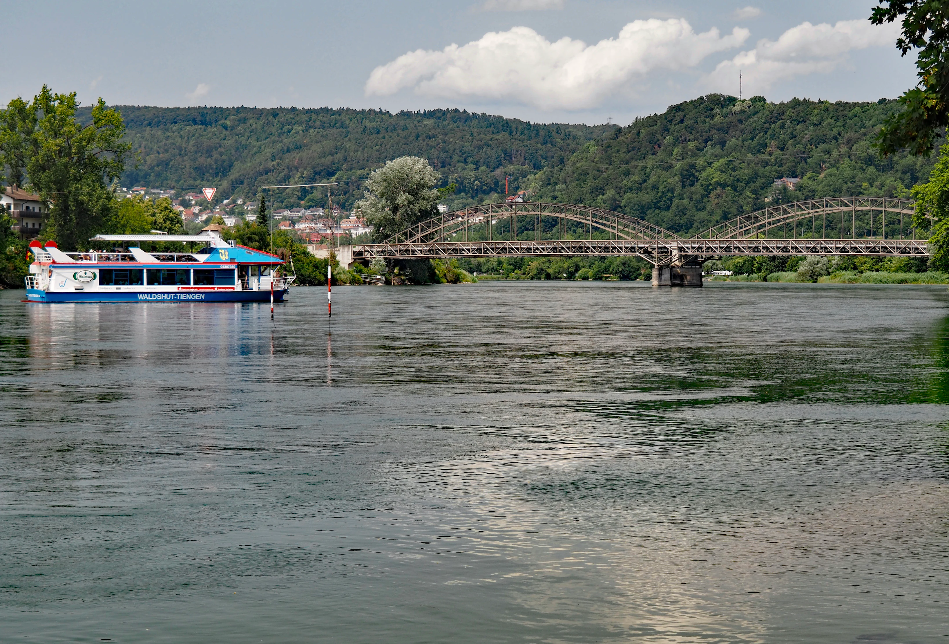 Un barco de pasajeros azul y blanco navega por el río Aare hacia un gran puente de celosía de acero en la distancia, con colinas boscosas al fondo.