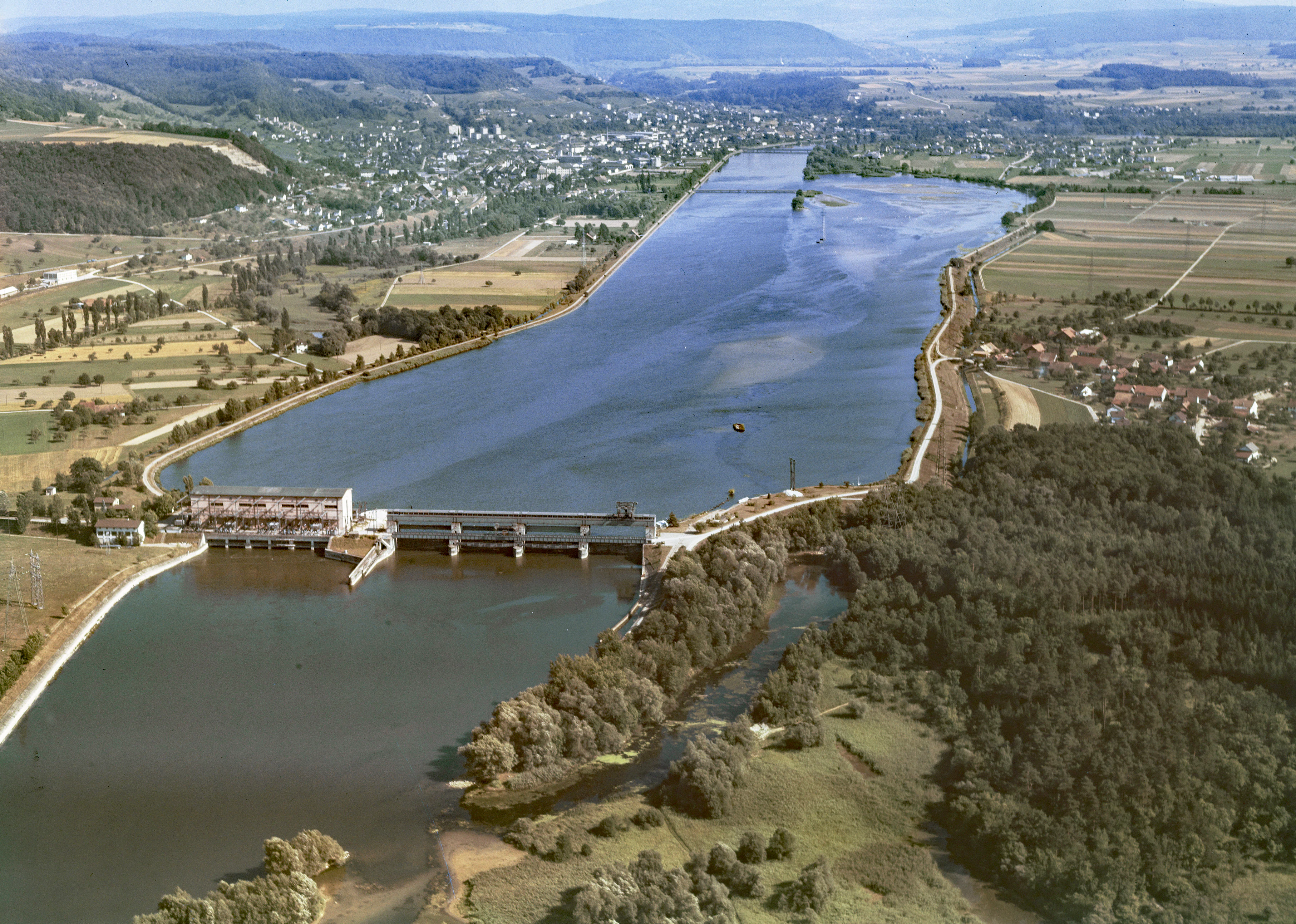 Fotografía aérea de gran angular del embalse de Klingnauer Stausee con su estructura de presa visible, rodeado de paisaje rural, bosques y campos en Argovia, Suiza.