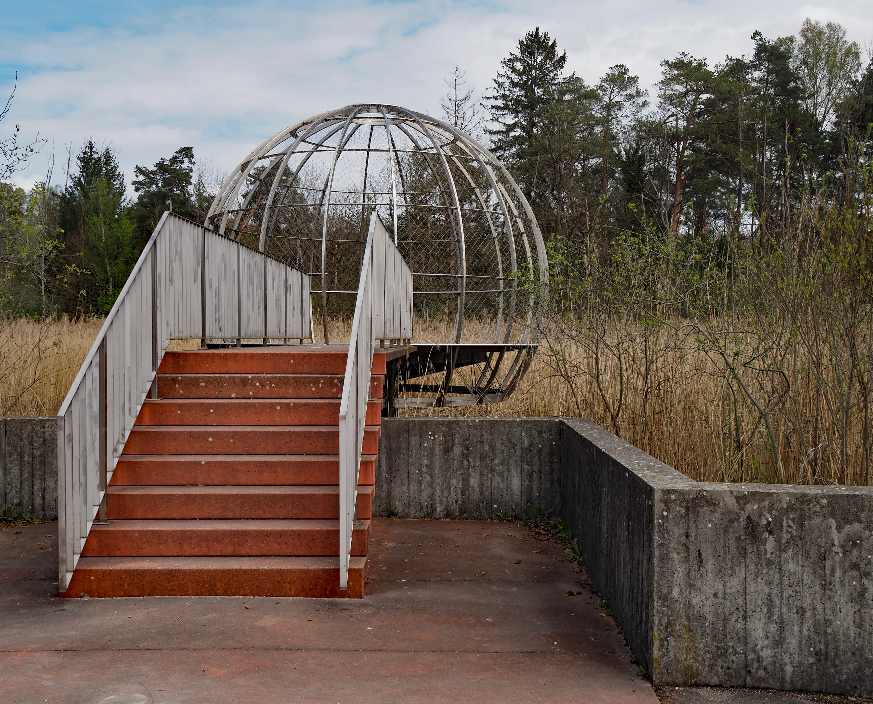 A modern spherical metal viewing platform structure at the top of a short set of stairs, set against a backdrop of tall grass and a pine forest.