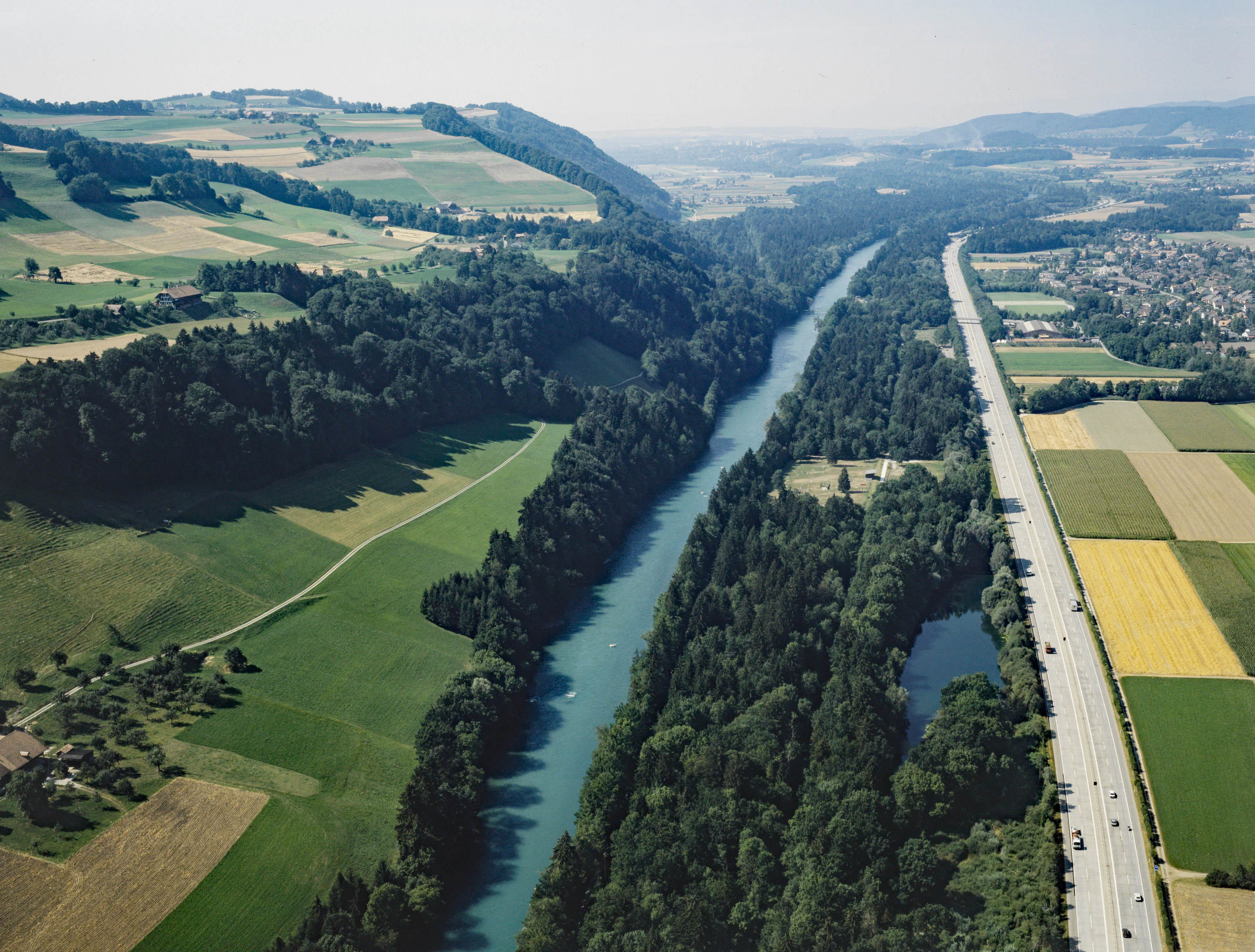 An aerial view showing the Aare River flowing through a lush green valley with the forested Belpberg hill on the left and a motorway running parallel on the right.