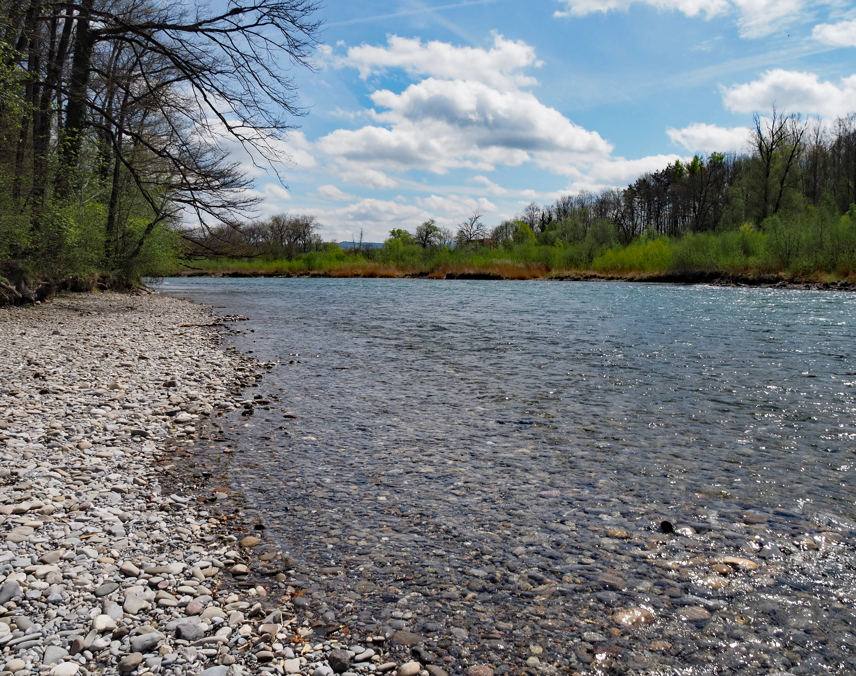 A clear, flowing river with a wide pebble and rock bank on the left; trees and greenery line the opposite riverbank under a blue sky with white clouds.