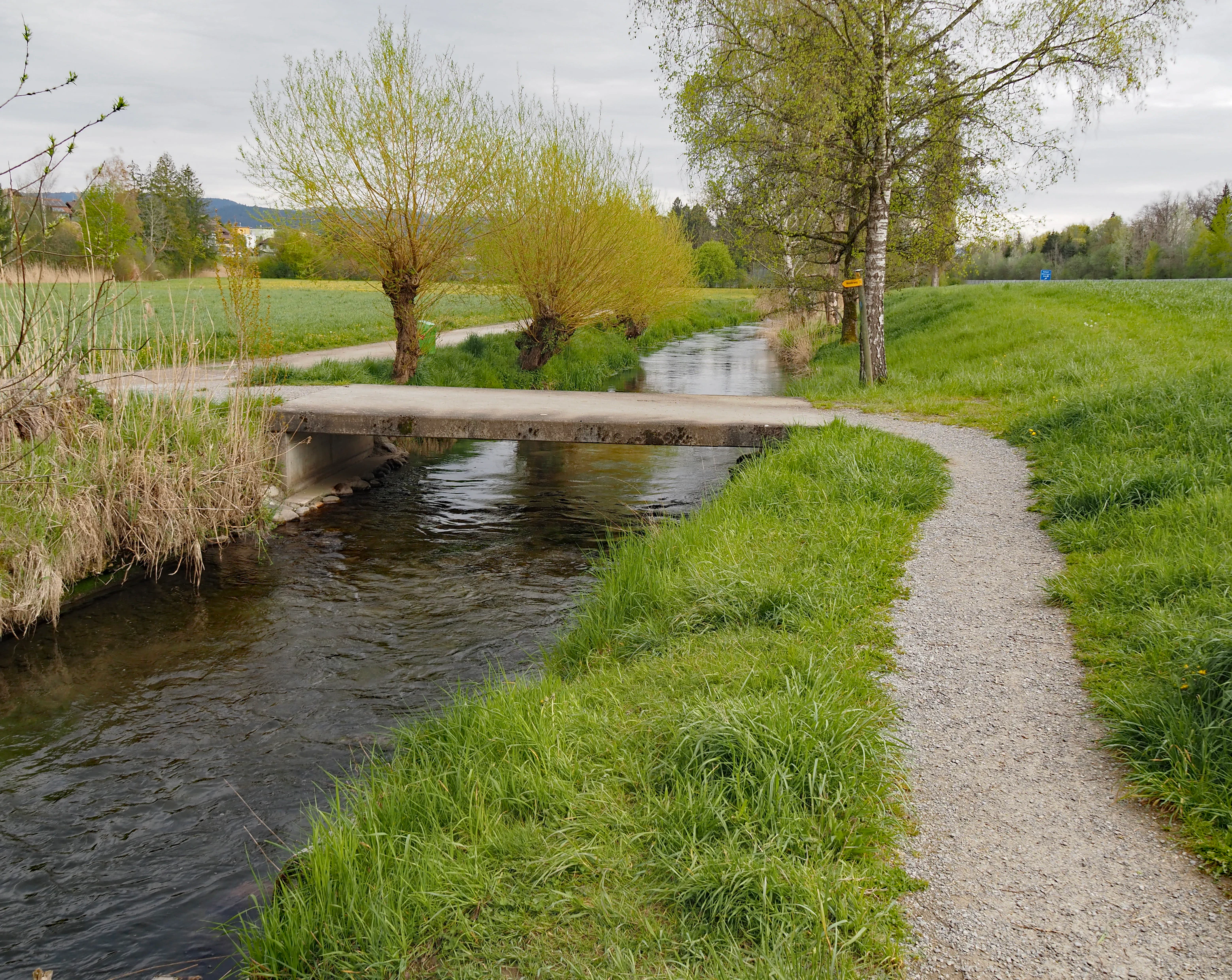 A narrow gravel walking path runs alongside a small stream; a simple concrete bridge crosses the water near several willow trees in a grassy landscape.