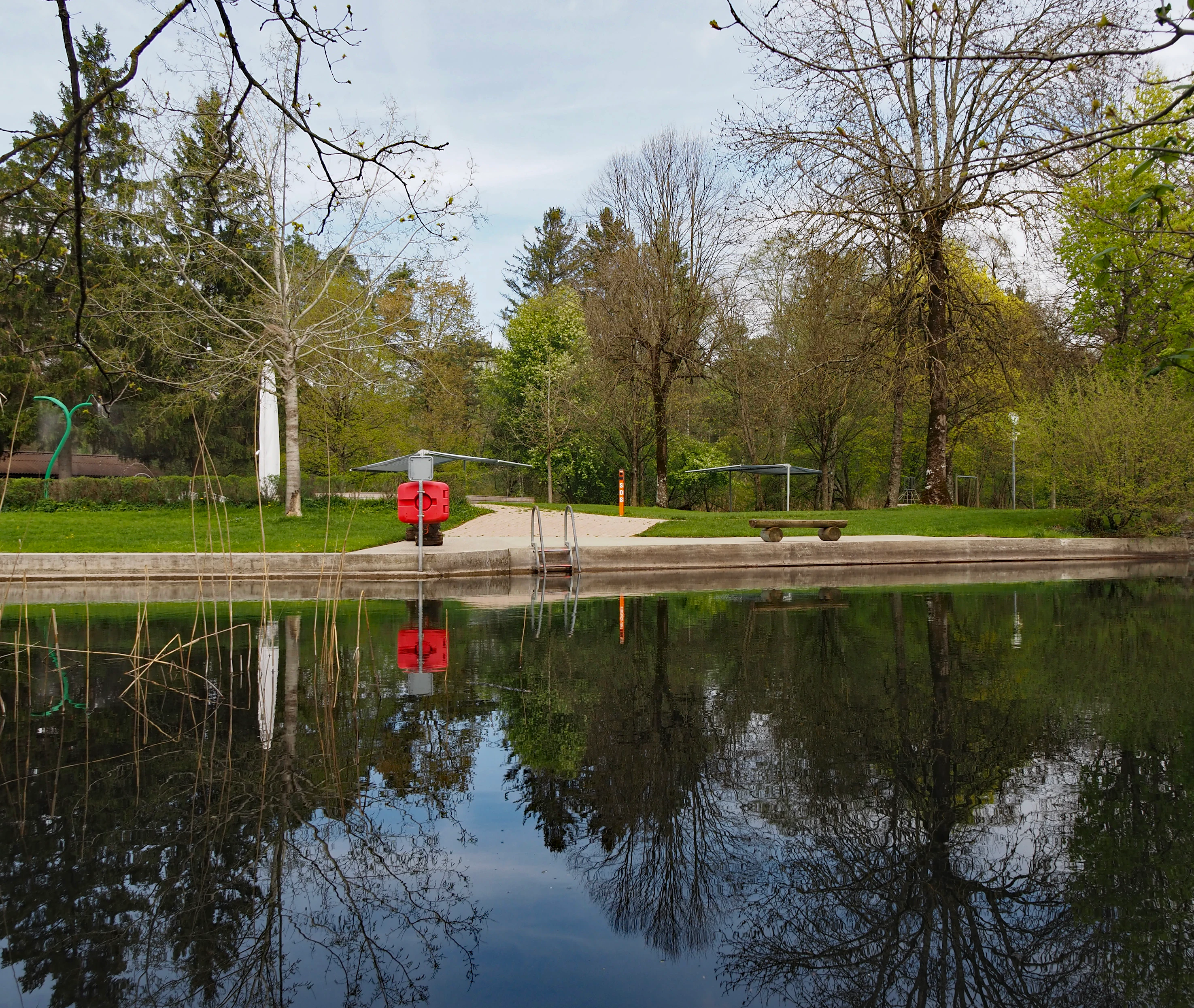 A calm, reflective river surface in the foreground, with a grassy bank, a red life ring, a wooden bench, and mature trees in the background at the Giessenbad swimming area.