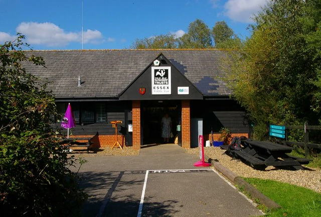 A single-story building with dark siding and a grey tiled roof serving as a visitor centre, featuring a gable entrance with a visible Essex Wildlife Trust sign.