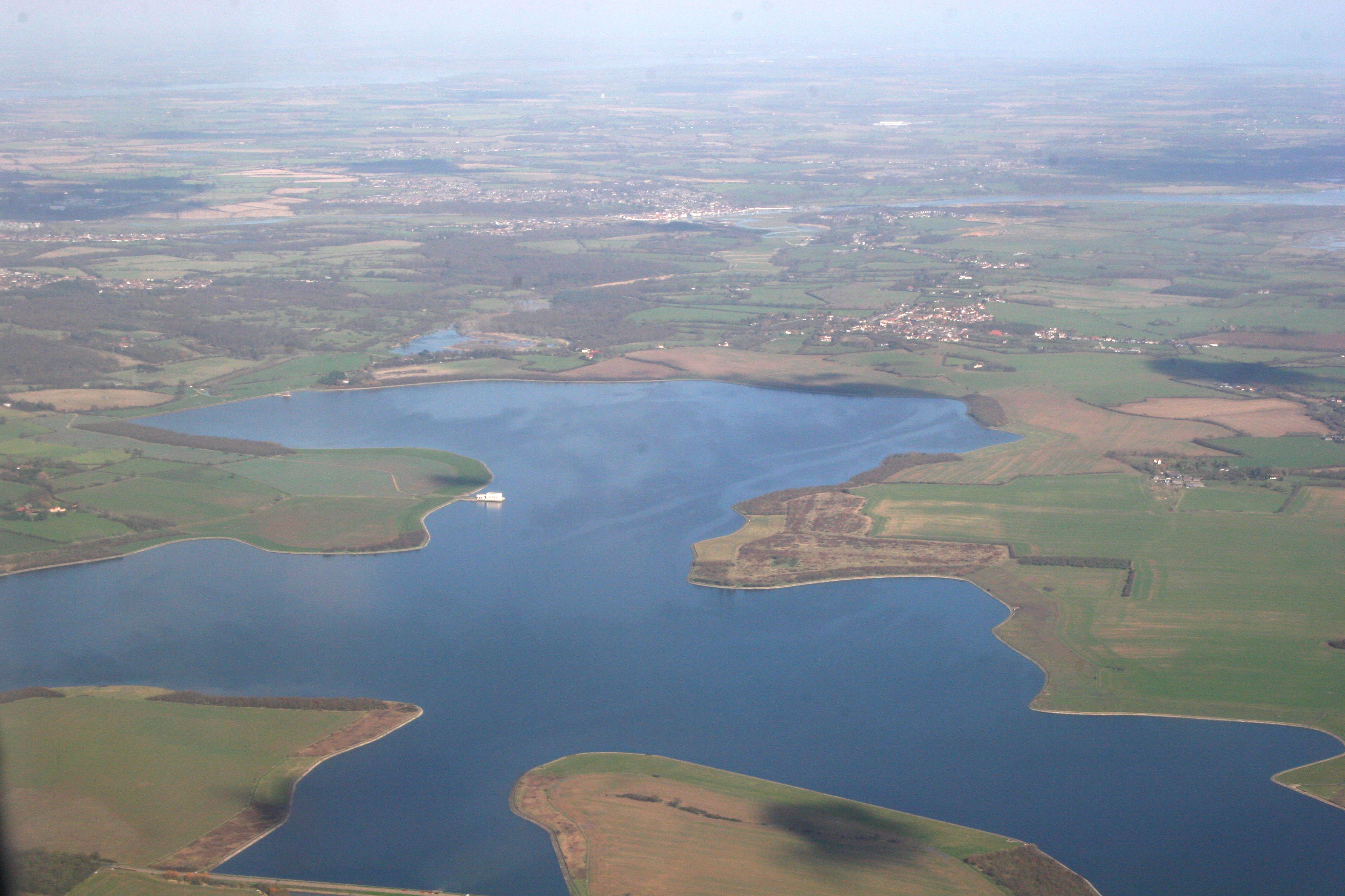 An aerial view of the Abberton Reservoir showing the expansive blue body of water surrounded by green and brown agricultural fields.
