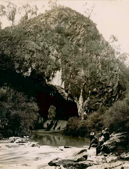 A vintage sepia-toned photograph showing a large limestone archway opening at the base of a rocky, tree-covered hillside, with a stream in the foreground and a group of people sitting on rocks nearby.