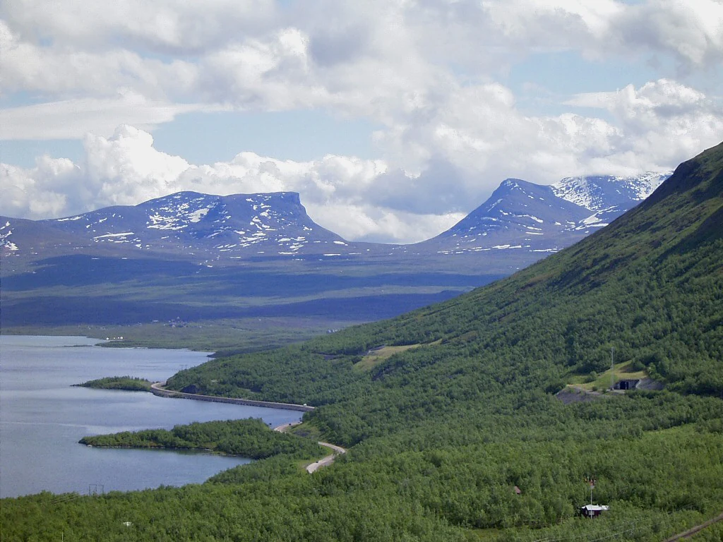 Landscape view of Lapporten valley with Torneträsk lake, snow-capped mountains, and dense green forest under a partly cloudy sky
