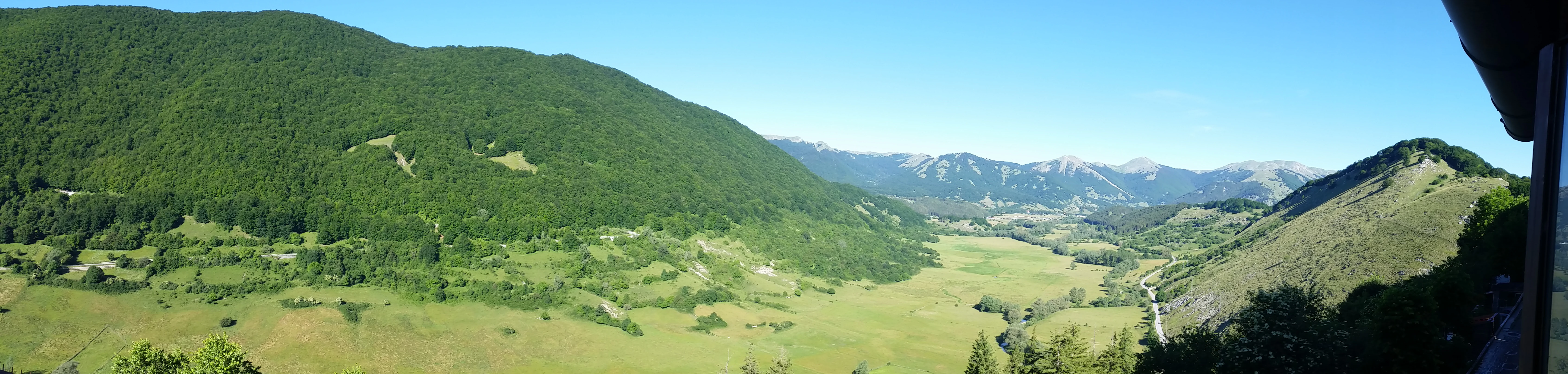 Panoramic landscape showing green mountains, valleys, and distant peaks under clear blue sky