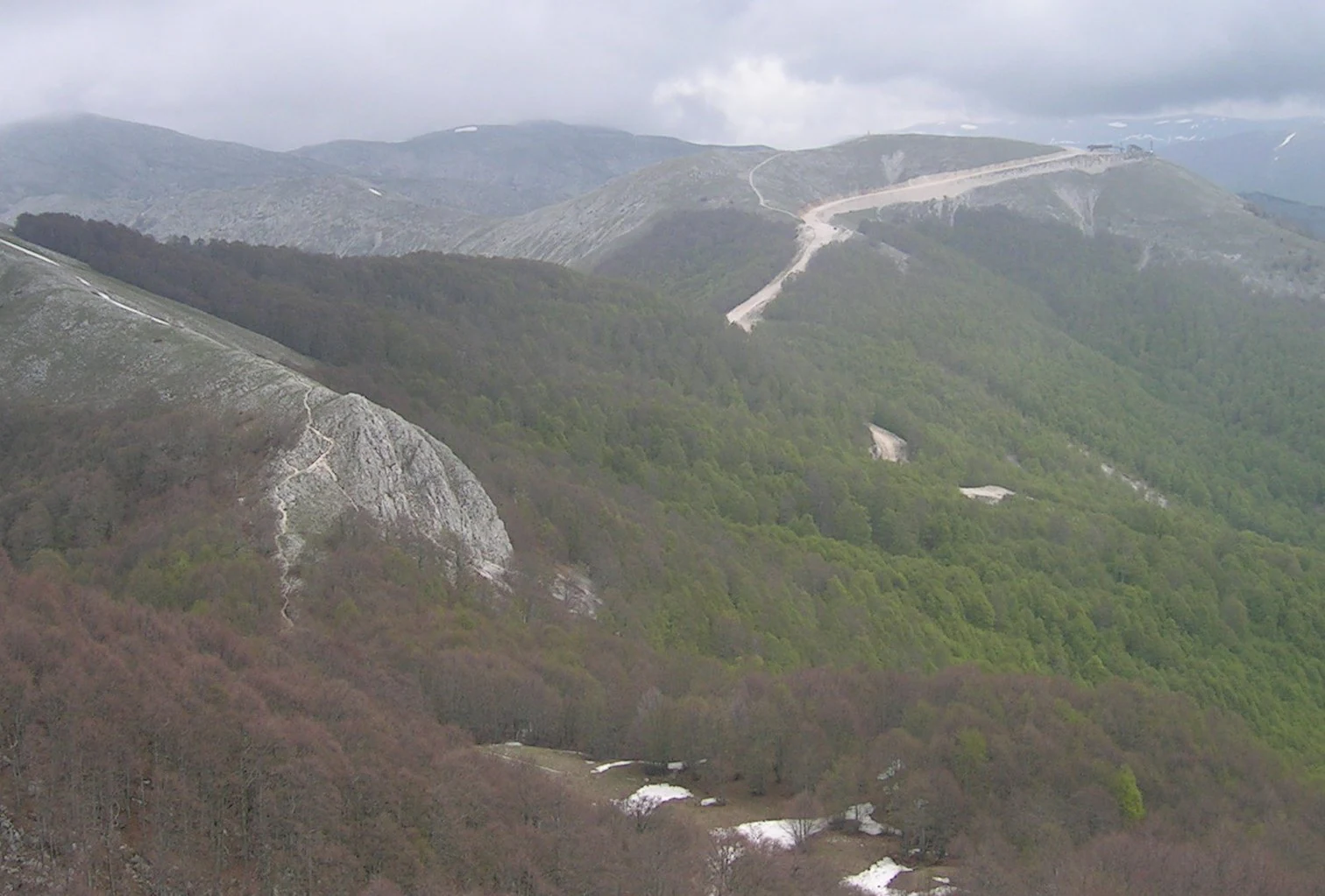 Panoramic mountain view with forested valleys, rocky outcrops, and a winding road under overcast sky