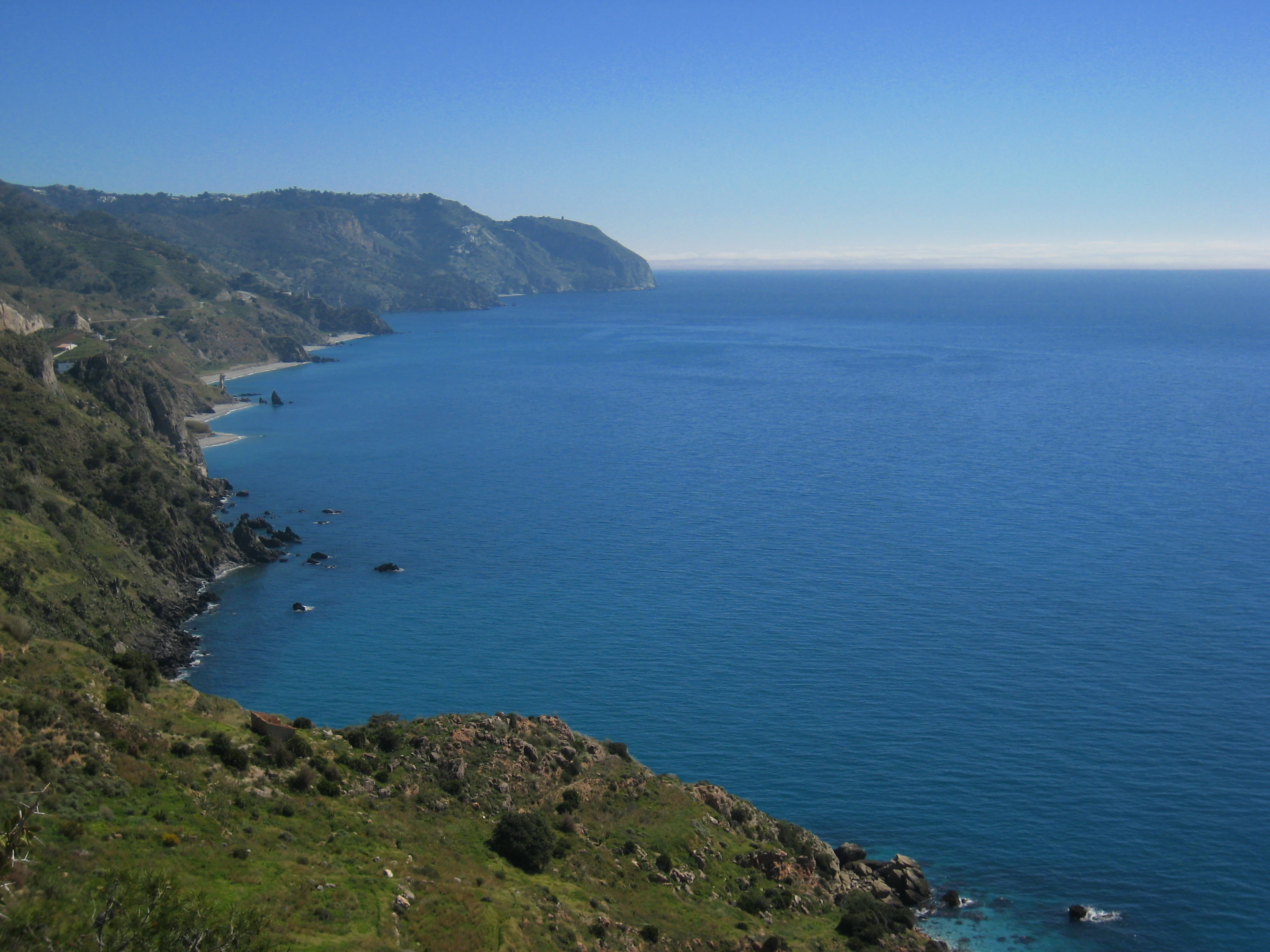 A wide-angle, high-angle view of steep, green-sloped cliffs dropping into the deep blue Mediterranean Sea along a sunny coastline.