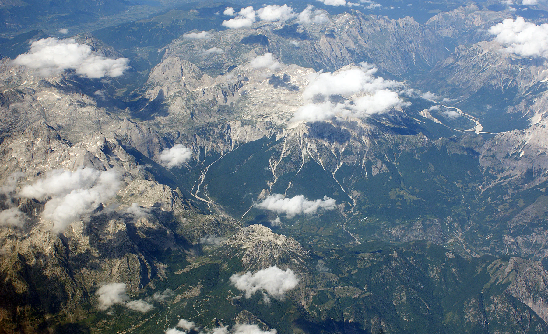 A high-altitude aerial view of the Accursed Mountains, showing jagged limestone peaks, forested valleys, and scattered clouds.