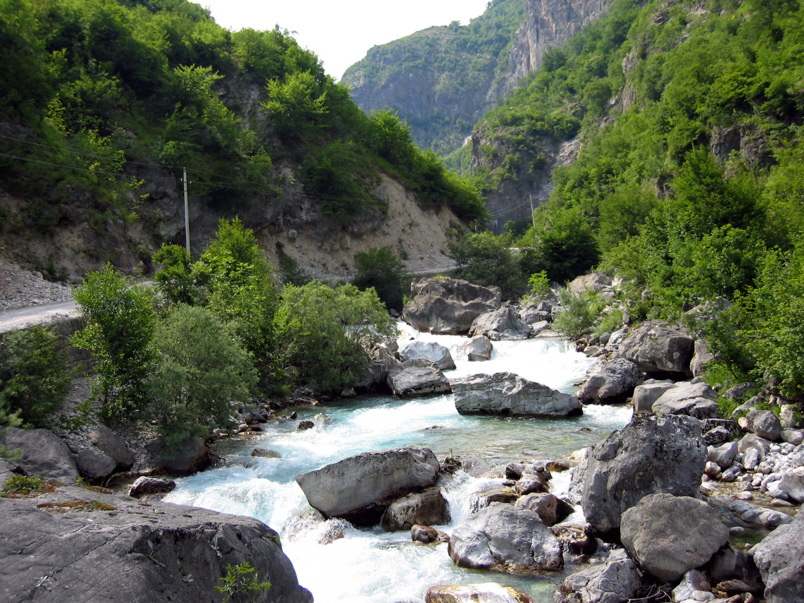 A rushing river with turquoise water and large boulders flows through a steep, green mountain valley.