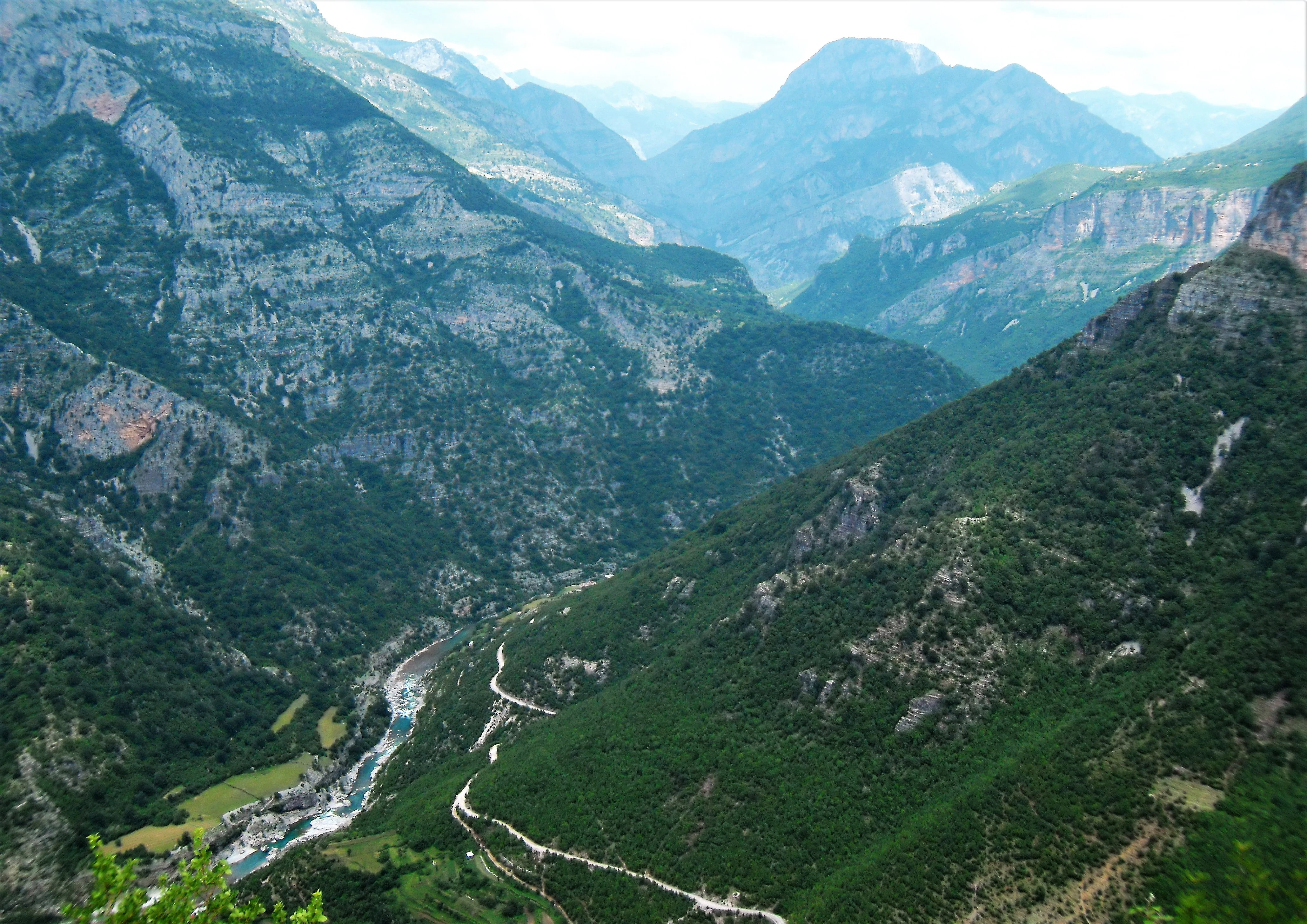 A high-angle scenic view of a deep, forested valley in the Accursed Mountains, featuring a river and a winding mountain road.