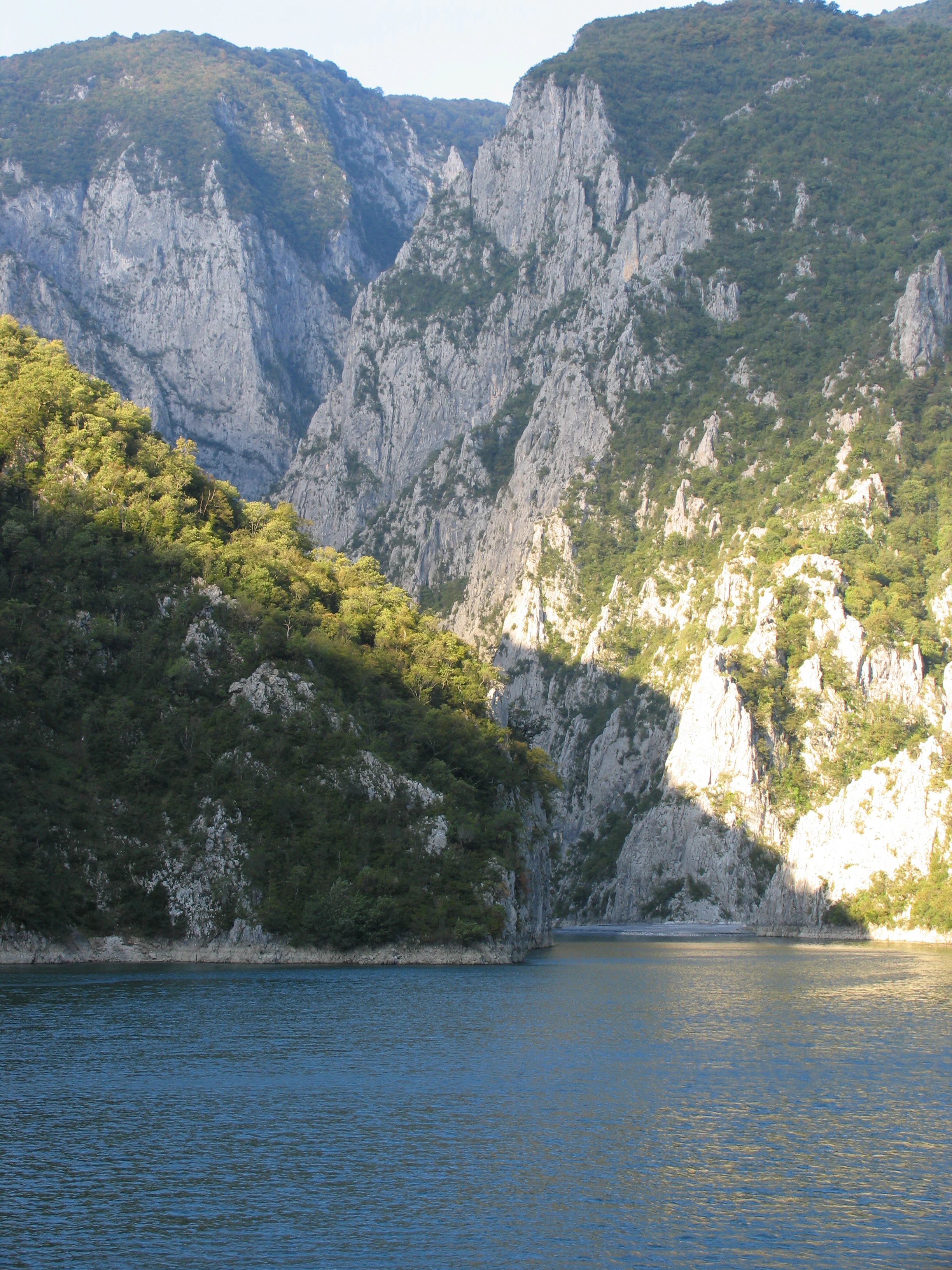 A view of the deep, calm waters of Lake Komani, surrounded by steep, craggy limestone mountains and dense forests in northern Albania.
