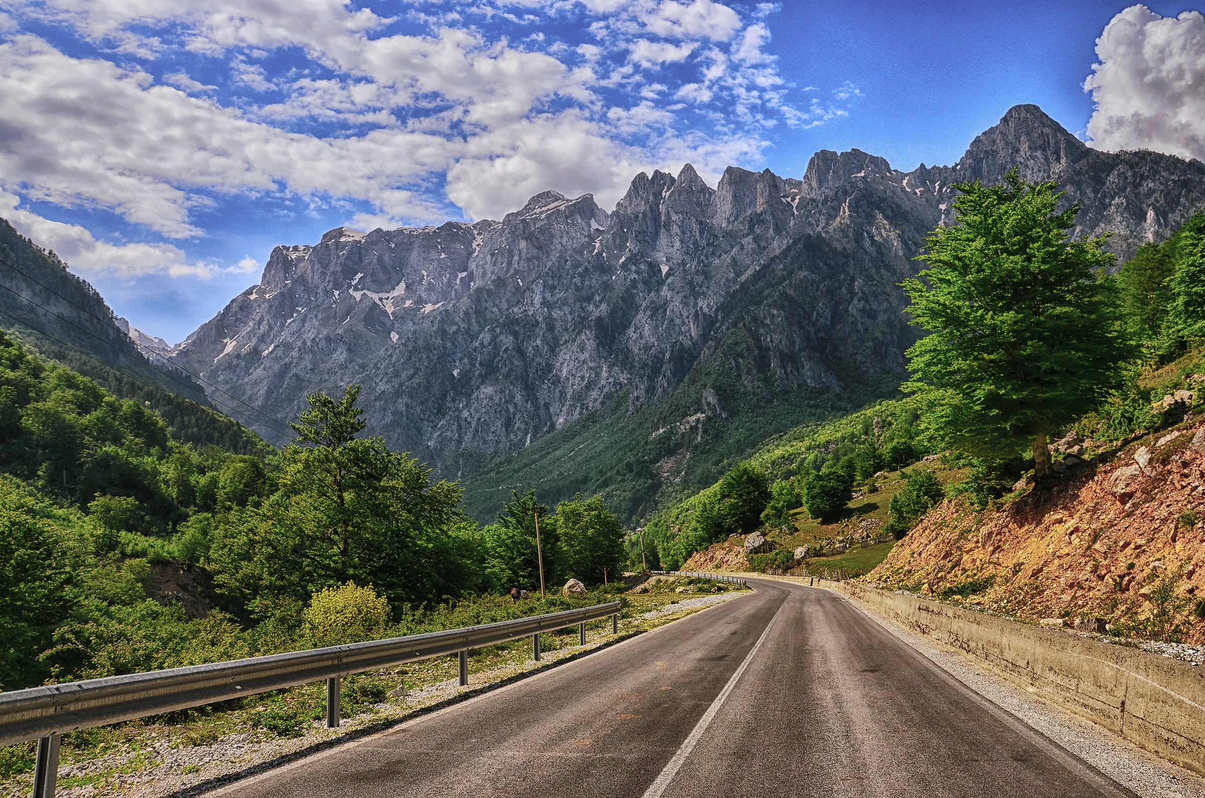 An asphalt road leads into a deep mountain valley flanked by lush green forests and towering, craggy limestone peaks under a blue sky with scattered clouds.