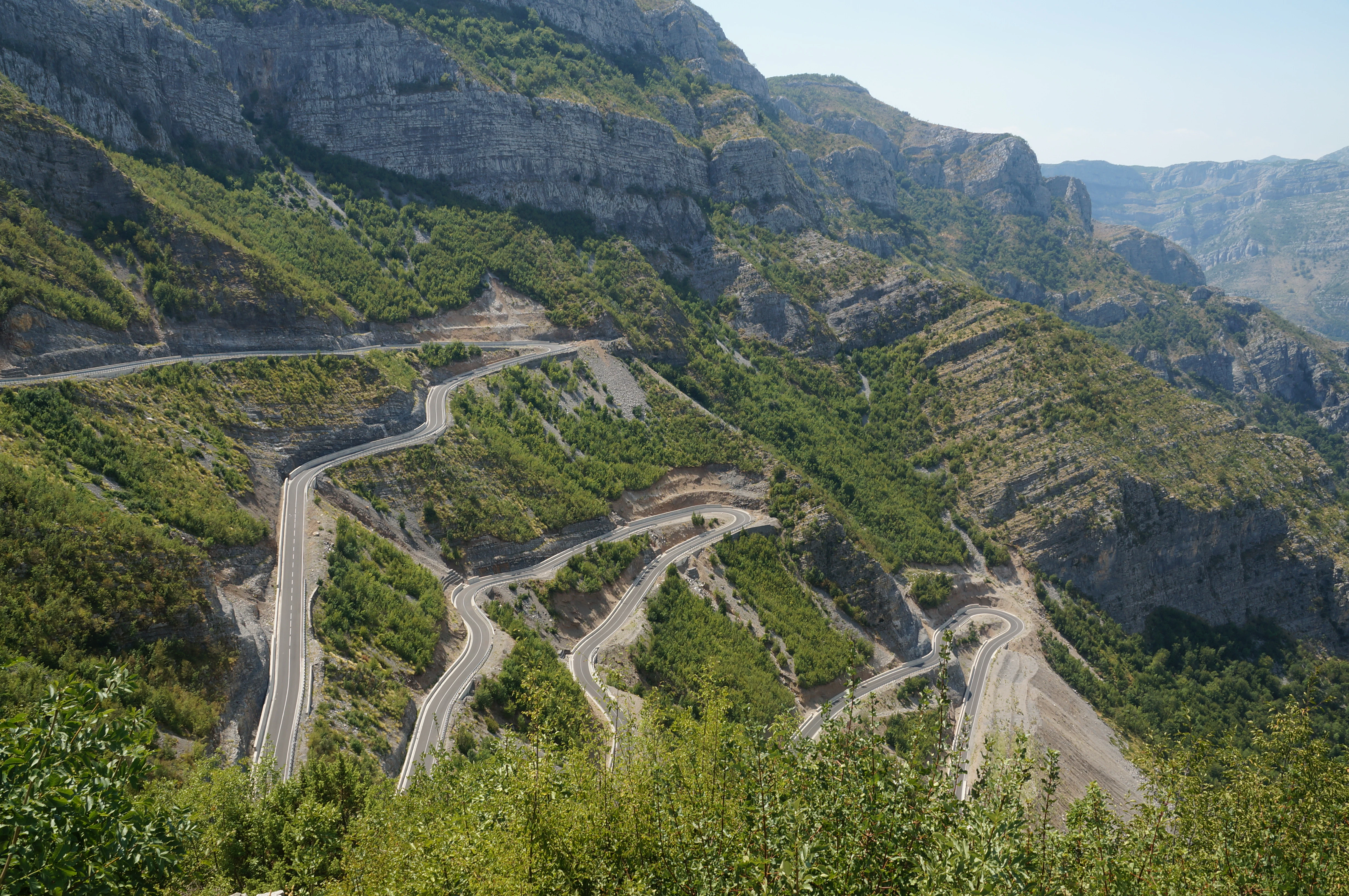 A steep mountainside with a paved road featuring multiple sharp switchbacks snaking down the slope, surrounded by rocky cliffs and green shrubs.