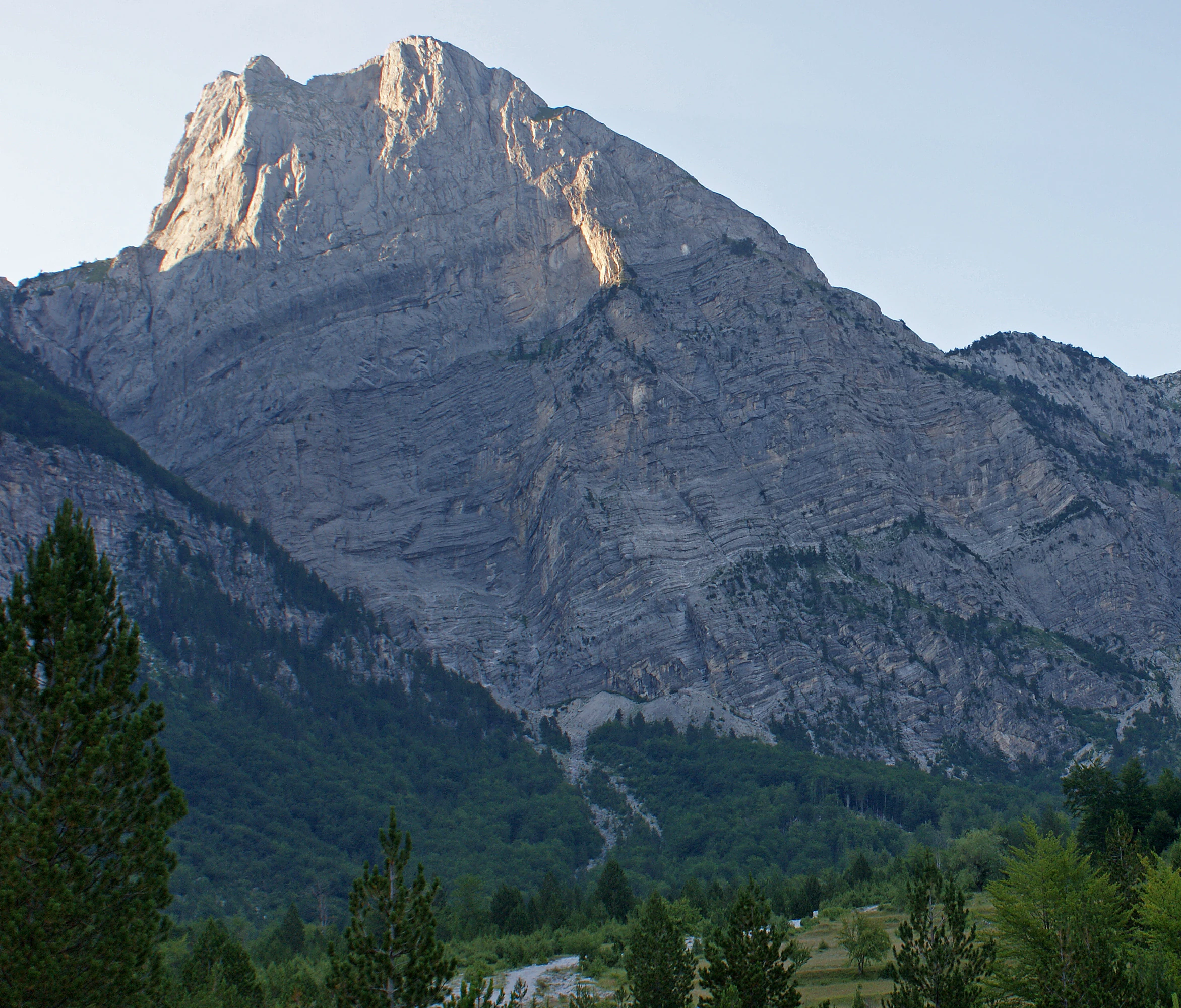 A towering rocky mountain peak rises above a lush green forest canopy in the Accursed Mountains.