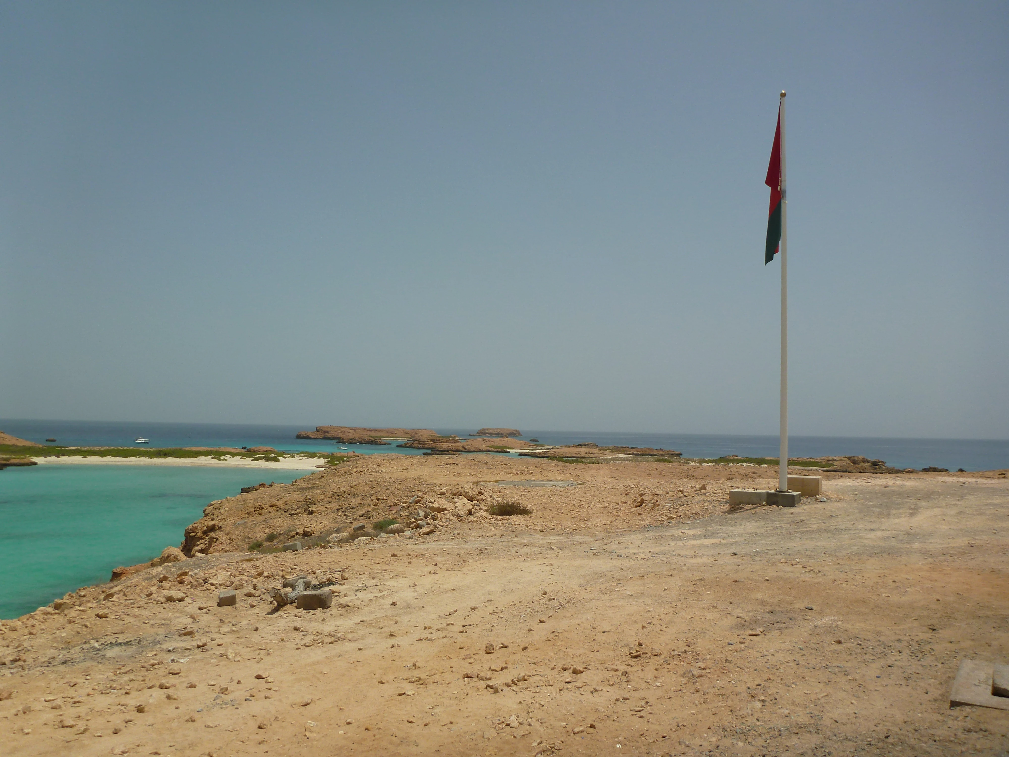 A wide shot of a rocky, arid island landscape surrounded by turquoise water, featuring an Omani flag on a white pole in the foreground.
