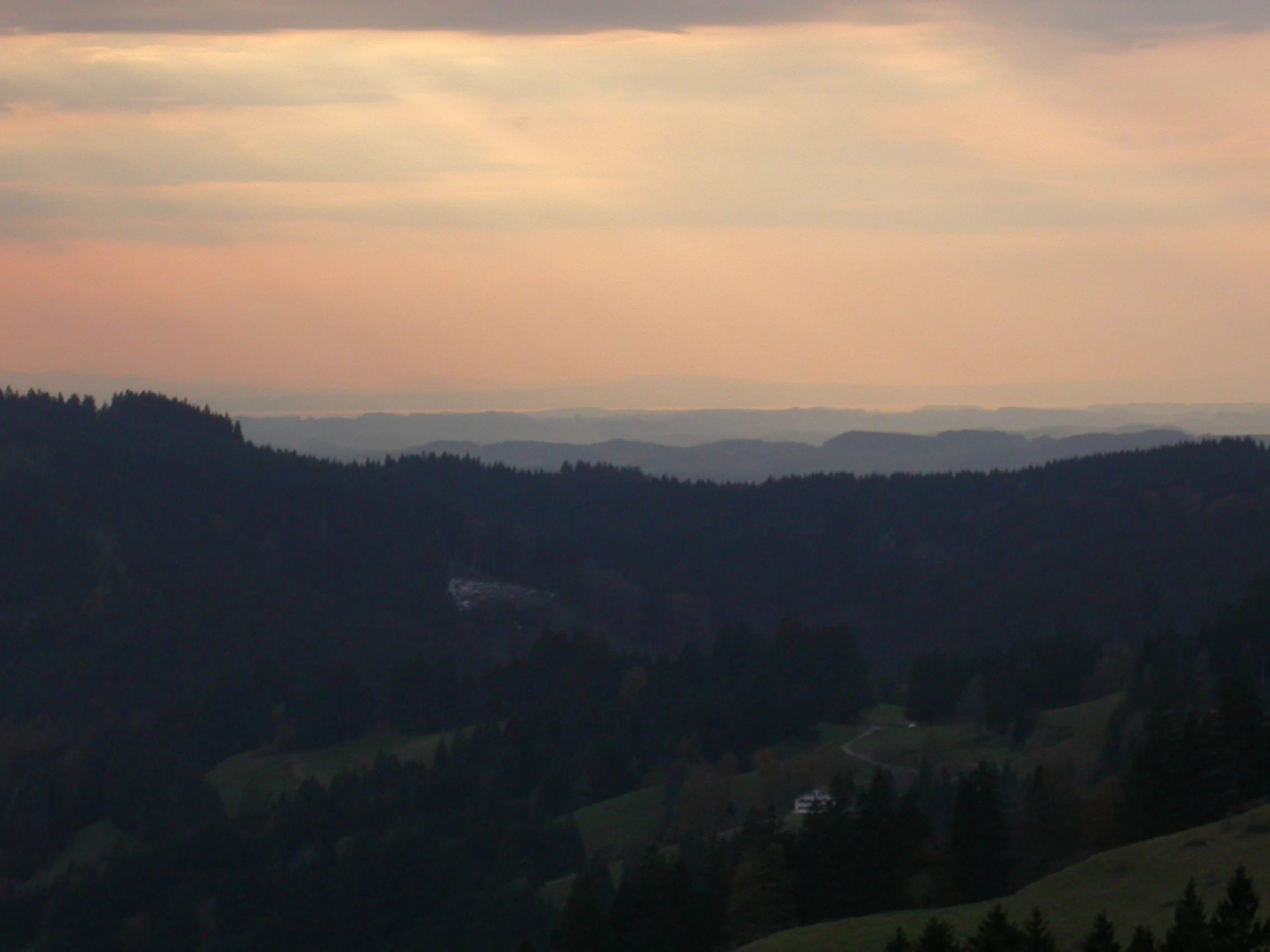 A wide, elevated landscape view featuring layers of forested ridgelines receding into a soft, hazy distance under a warm-toned sunset sky.