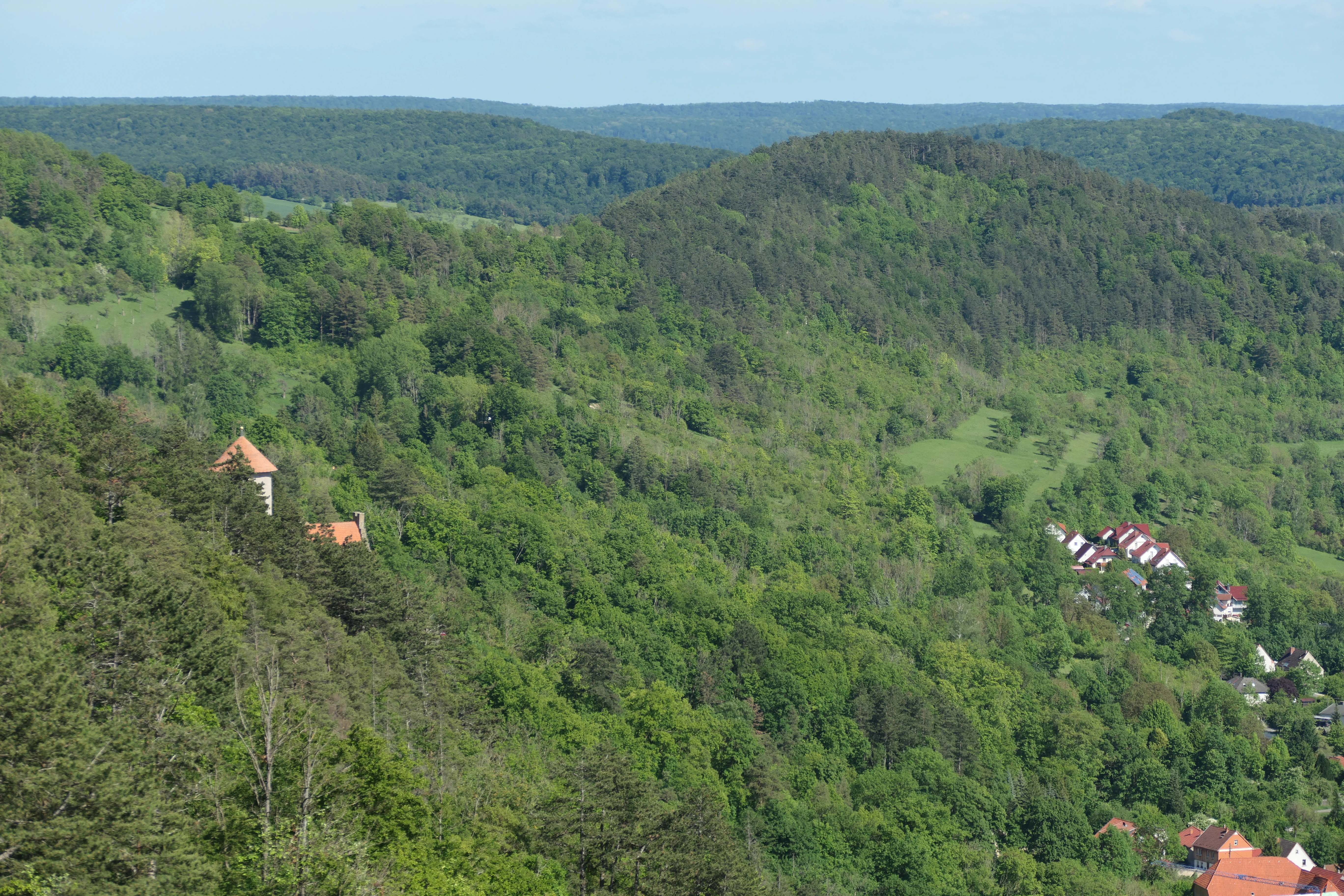 Un paisaje verde, montañoso y boscoso en Alemania con casas dispersas y un edificio prominente con techo de tejas rojas visible en la ladera izquierda.