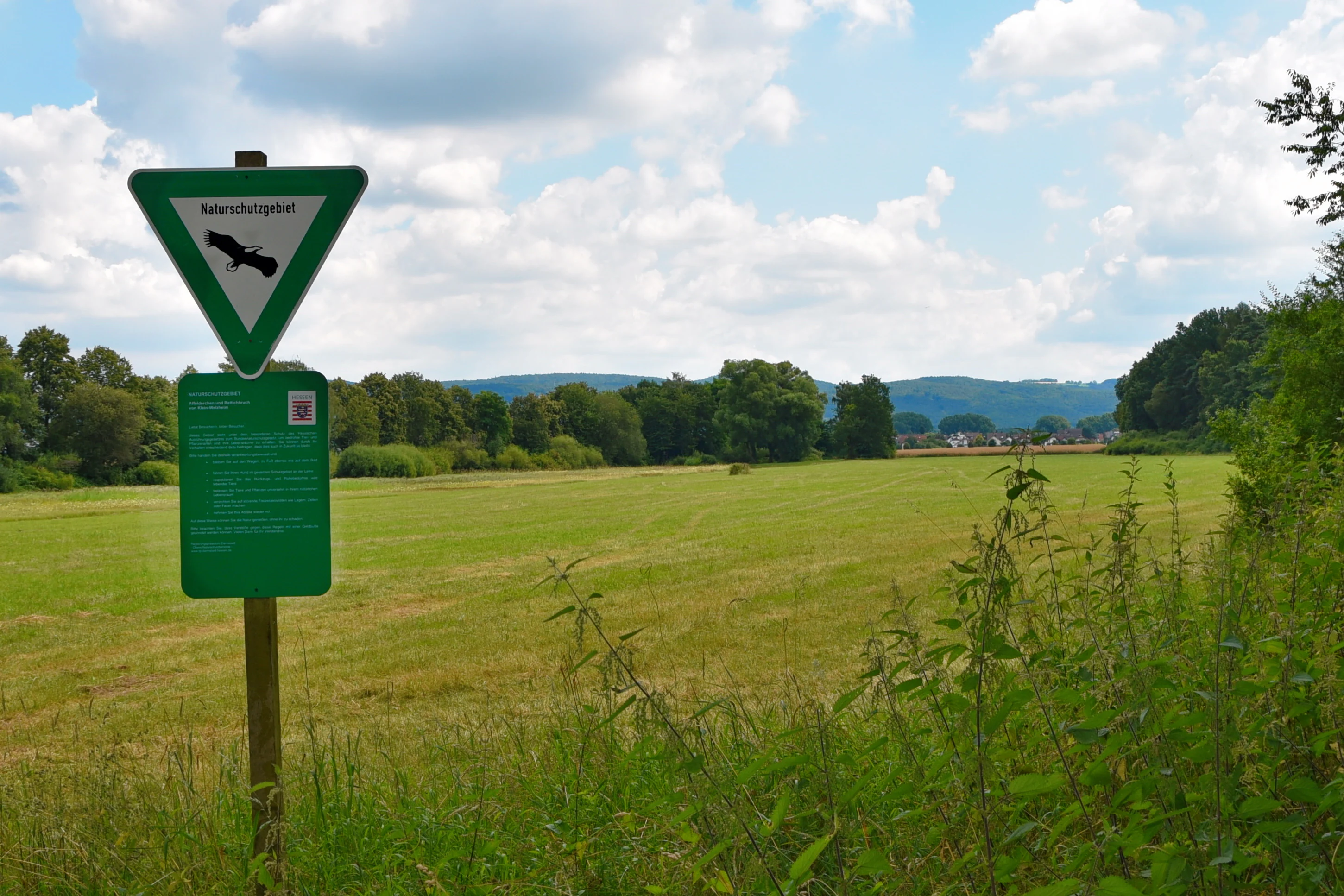 A green triangular nature reserve sign with a black bird icon stands on a wooden post in a grassy field in Germany.