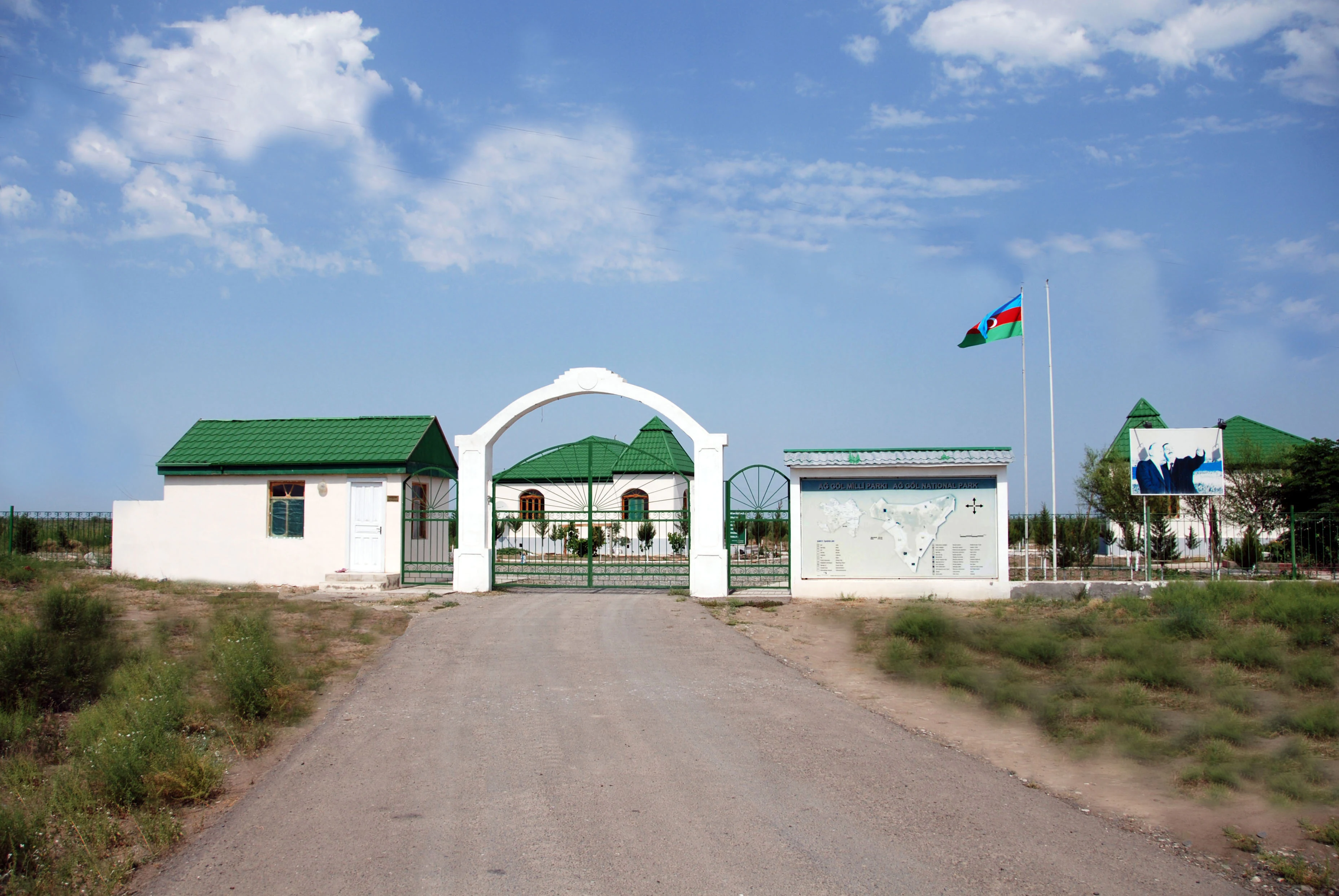 White archway entrance gate with green-roofed buildings on either side, dirt road leading to gate, and Azerbaijani flag flying in background.