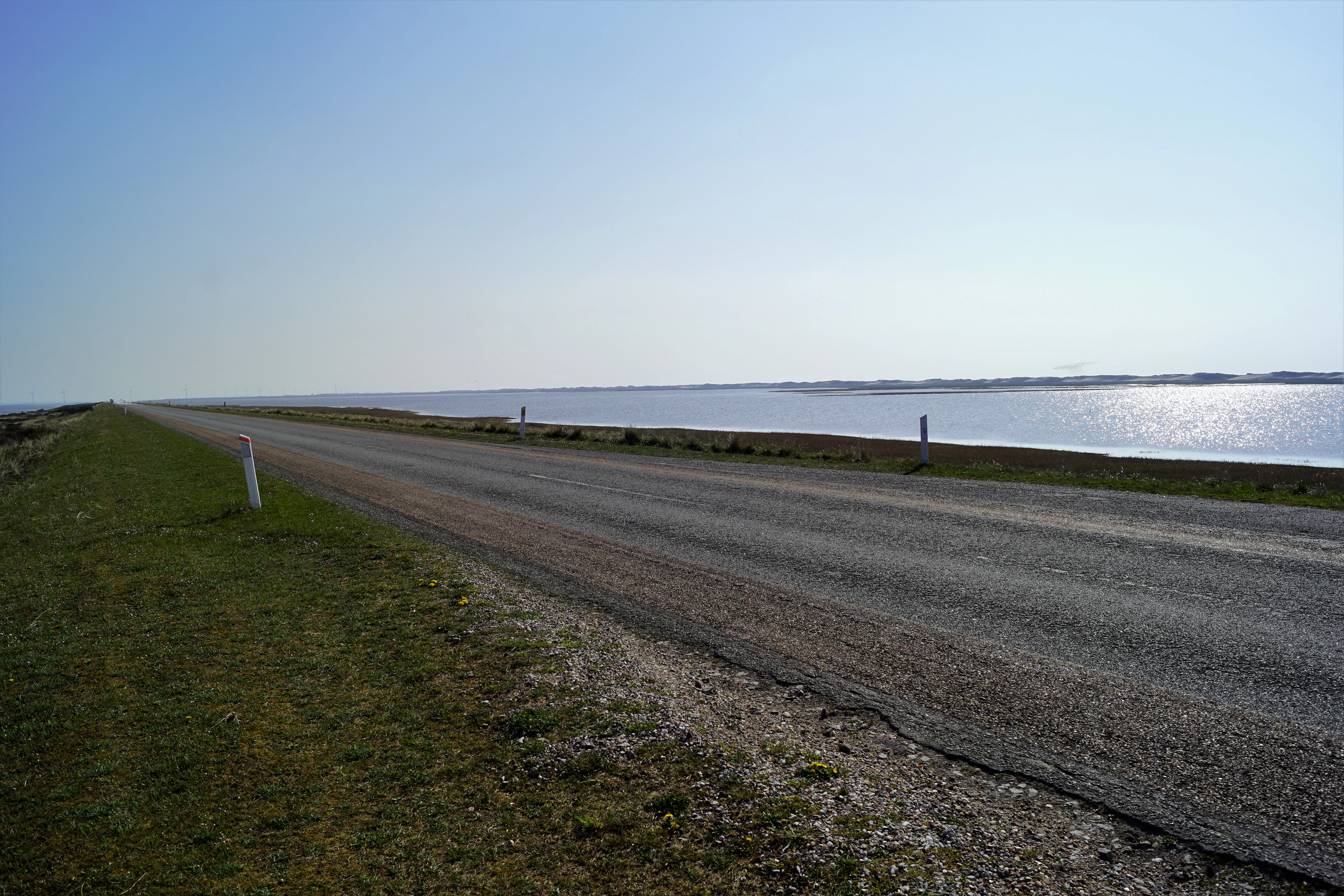 A paved road stretches into the distance, bordered by grassy banks and a calm expanse of water under a clear, bright sky.