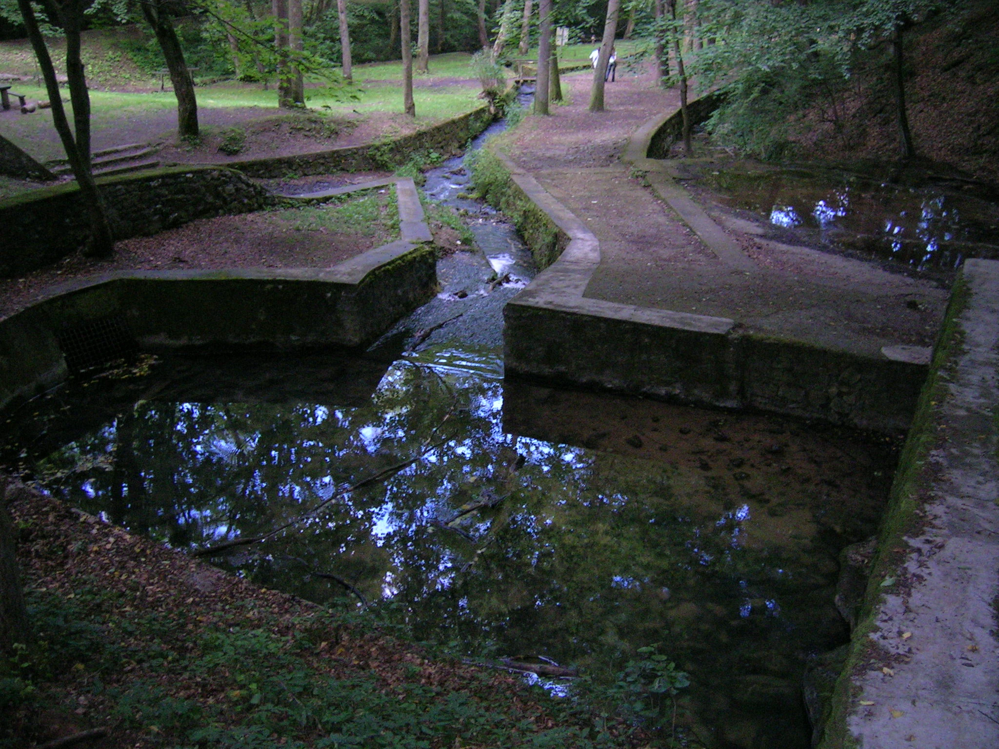 Small concrete-lined stream with reflective water surrounded by wooded area and pathway