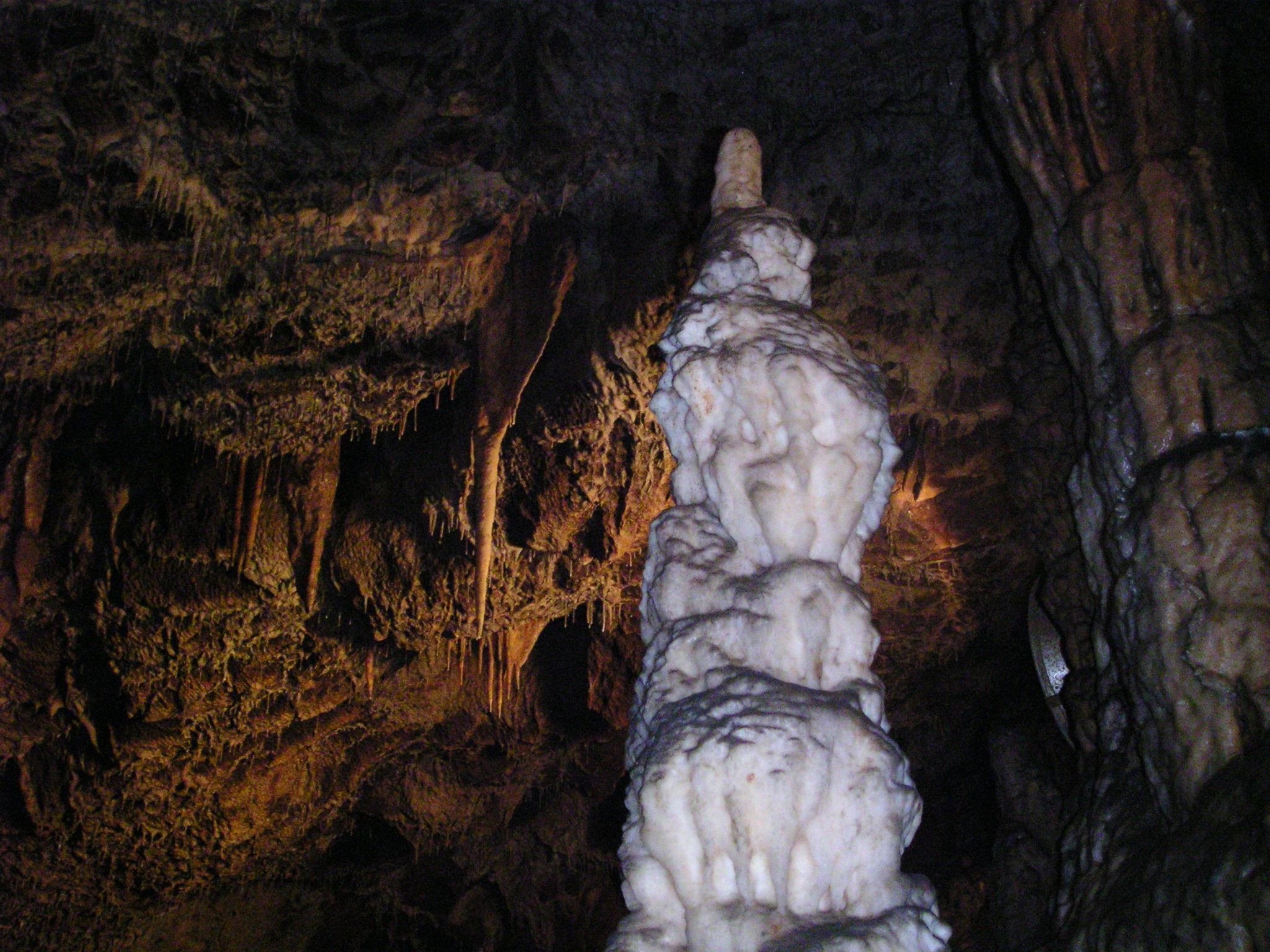 Tall white stalagmite in a dark cave with surrounding rock formations and dim artificial lighting