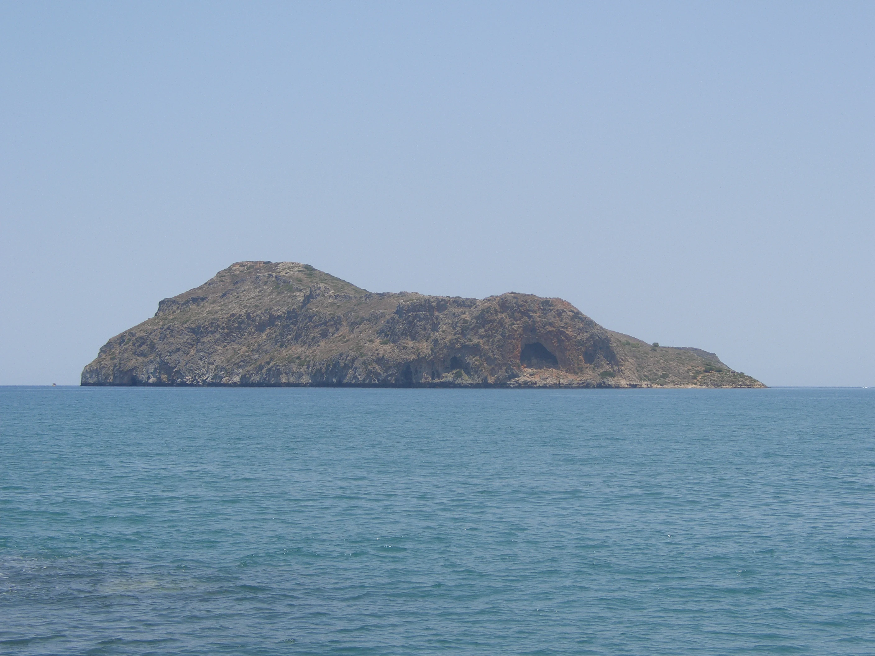 A wide, eye level shot of a small, barren, rocky island rising out of a calm blue sea under a clear sky.