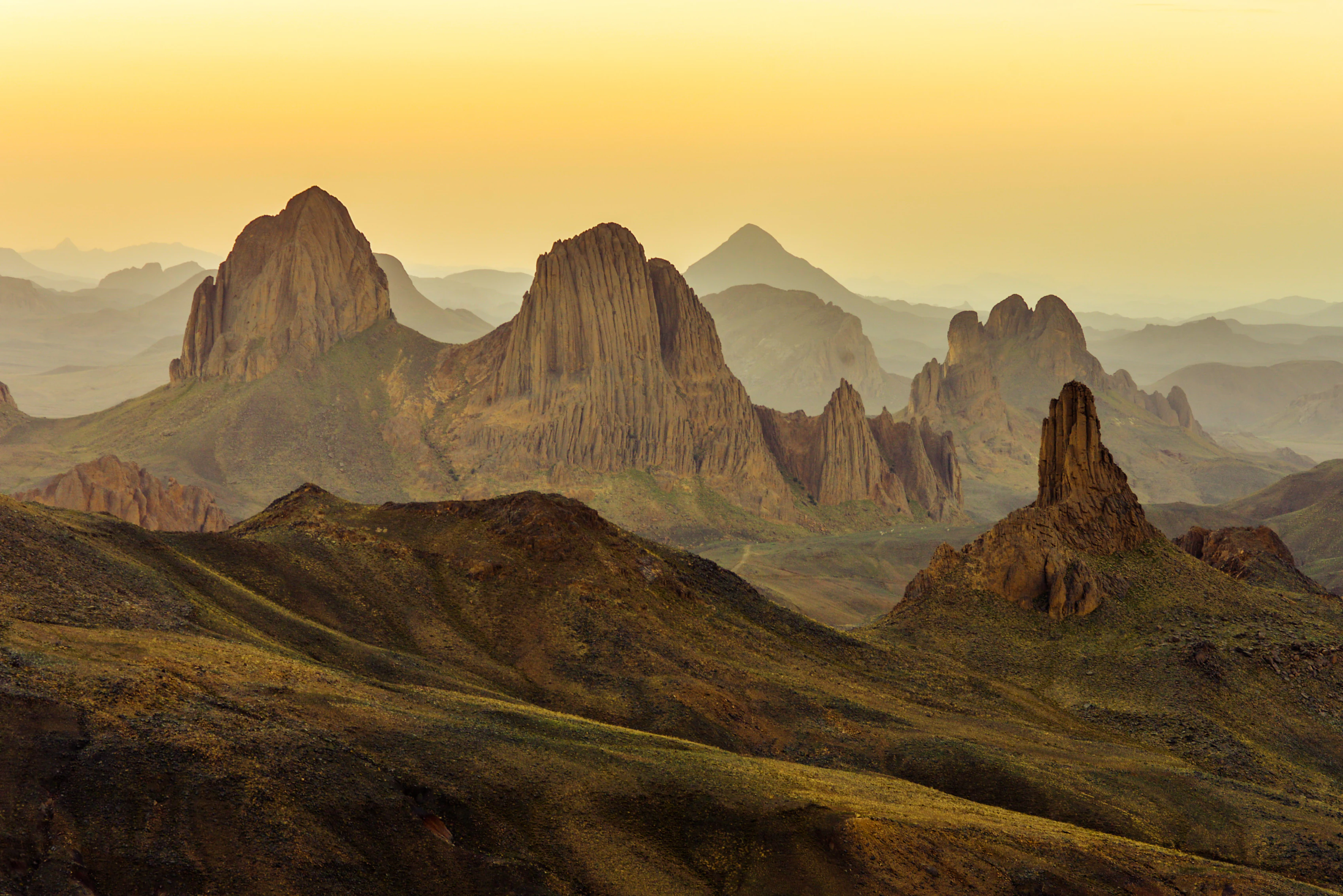 Wide angle photograph of the rugged Hoggar Mountains landscape in Algeria at sunset, featuring sharp volcanic peaks and deep valleys in warm golden lighting.