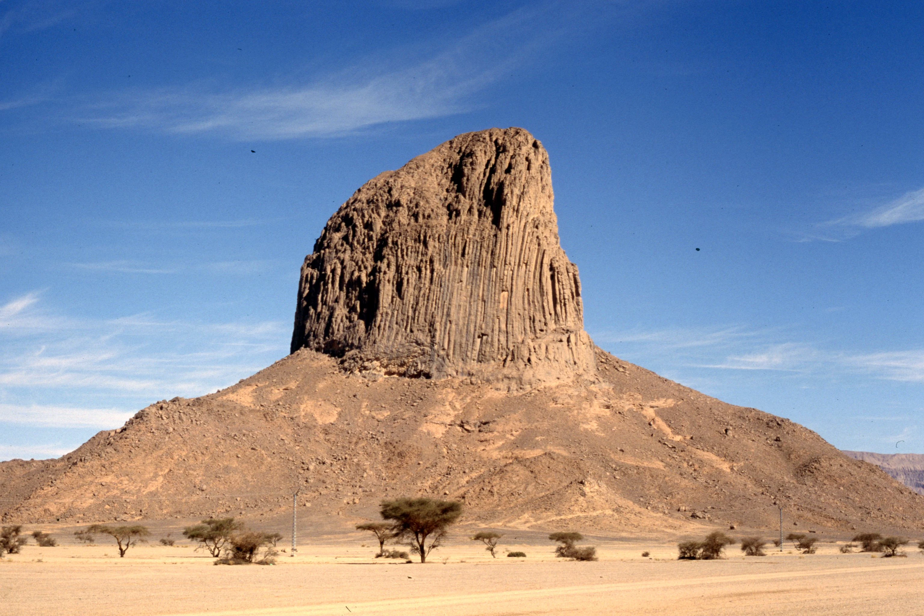 A large, isolated volcanic rock pinnacle rises above a sparse, arid desert landscape under a clear blue sky.