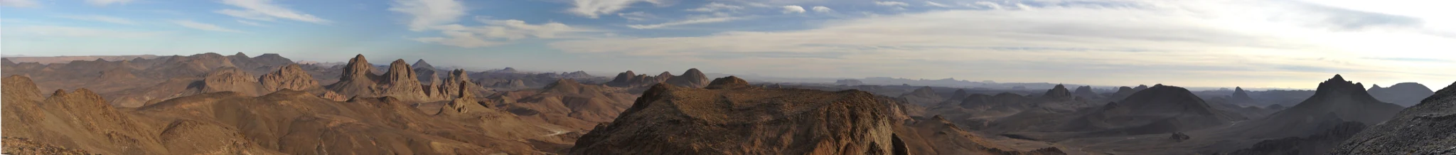 A wide-angle panoramic photograph showing a vast, arid mountain range with jagged rocky peaks and deep valleys under a light sky.