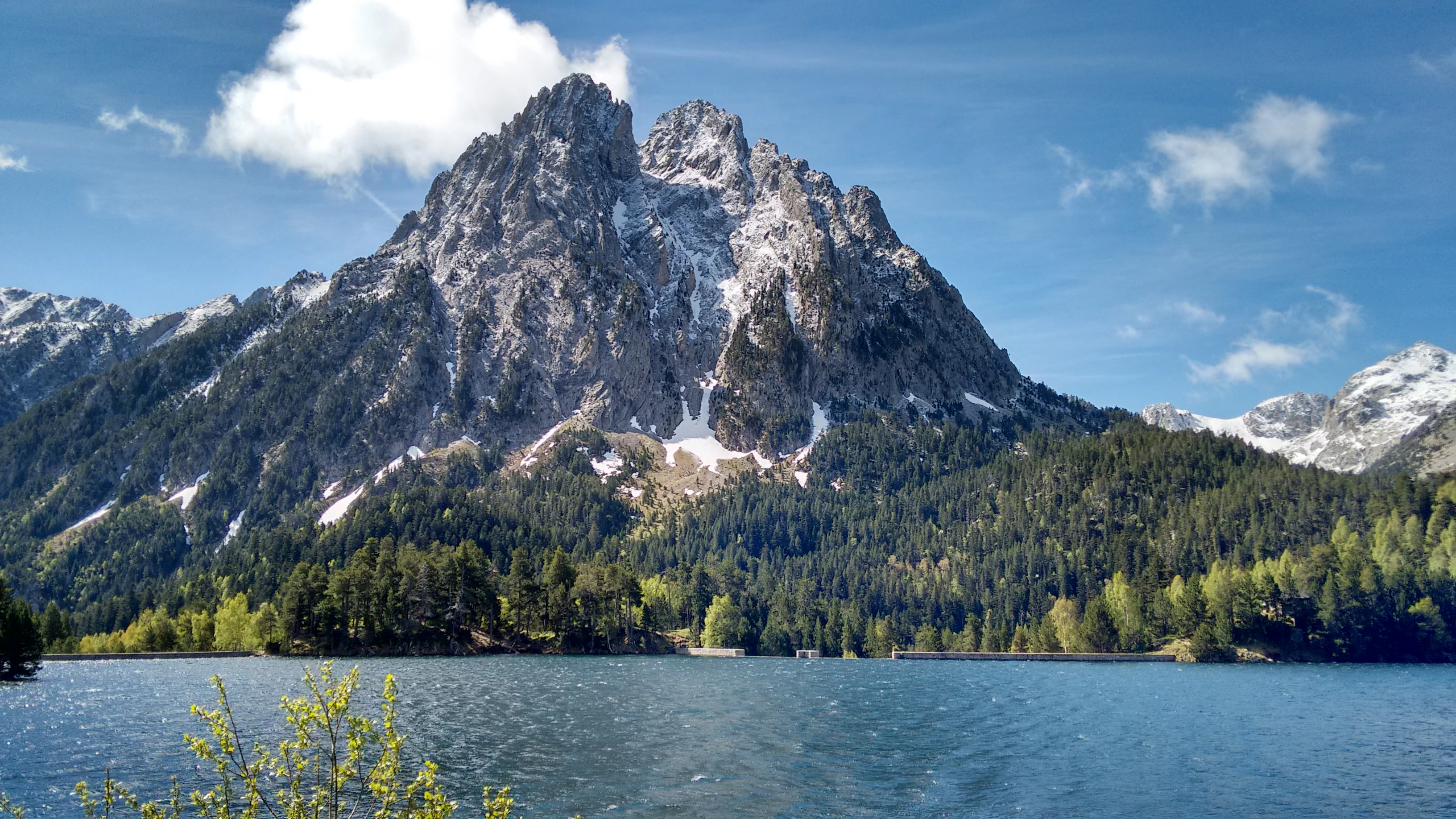 A mountain peak with rocky terrain and snow patches above a calm lake surrounded by forested hills under a blue sky with scattered clouds