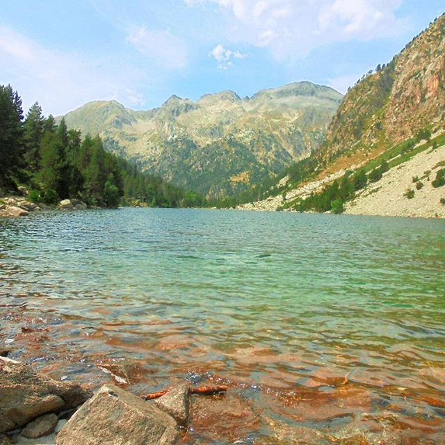 Glacial lake with clear turquoise water bordered by rocky shore and forested mountains under a partly cloudy sky.