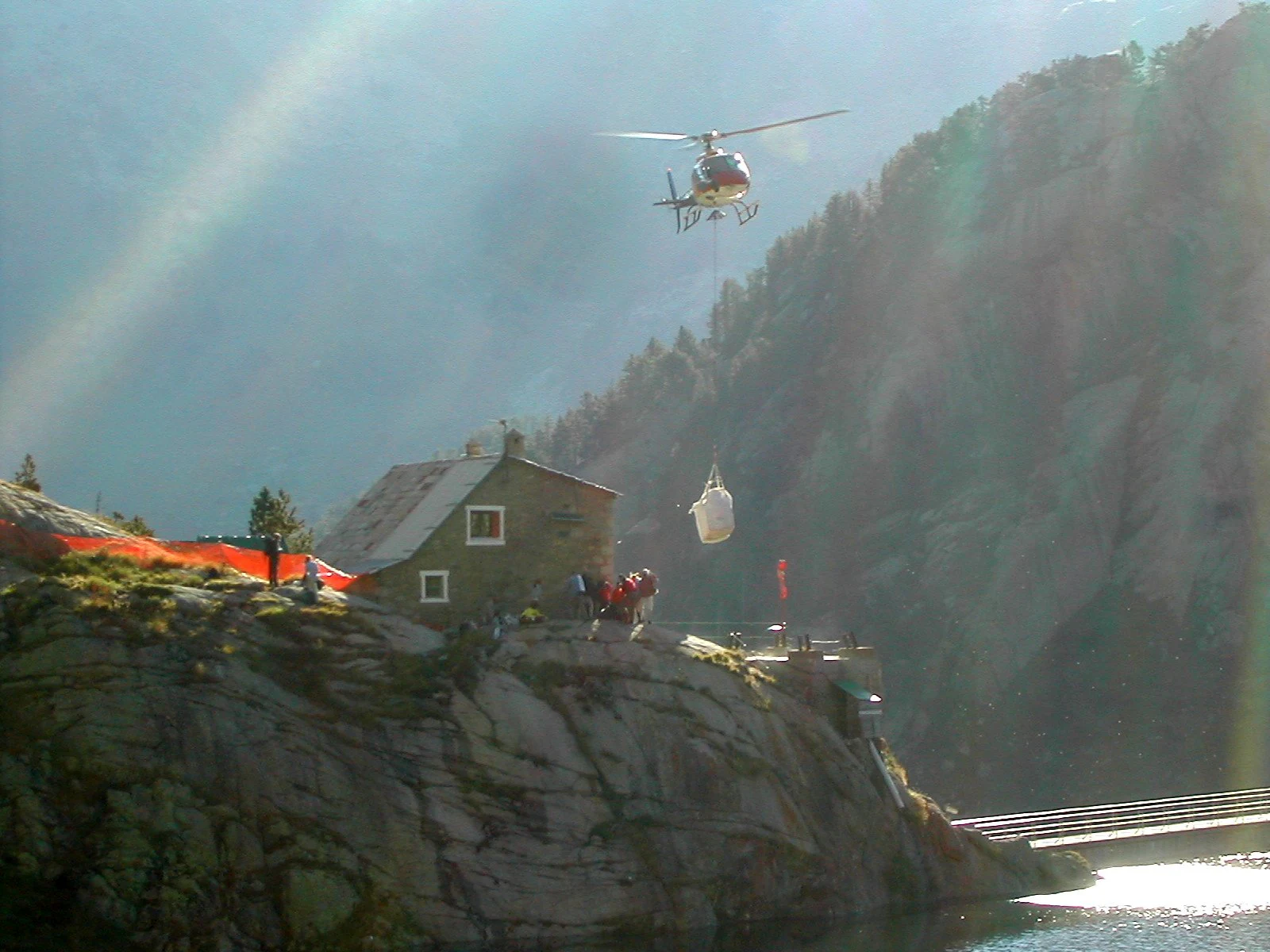A helicopter lowers a cargo container to a mountain refuge building on a rocky cliffside with a lake and forested mountains in the background