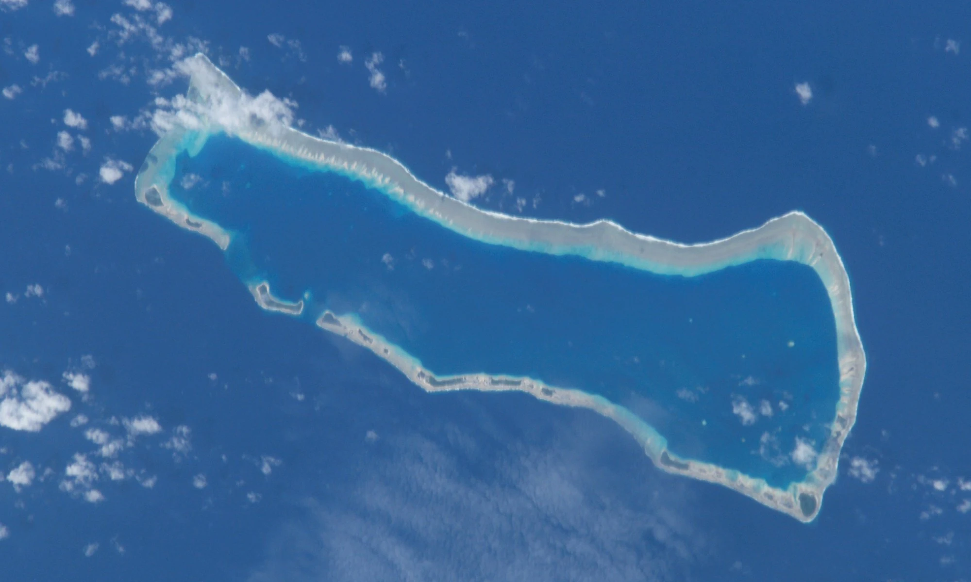 An overhead satellite image of Ailinginae Atoll showing a ring of coral islands and reefs enclosing a deep blue central lagoon, surrounded by the dark blue open ocean.