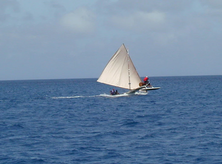 A small traditional sailing canoe with a triangular sail moving across deep blue open water.