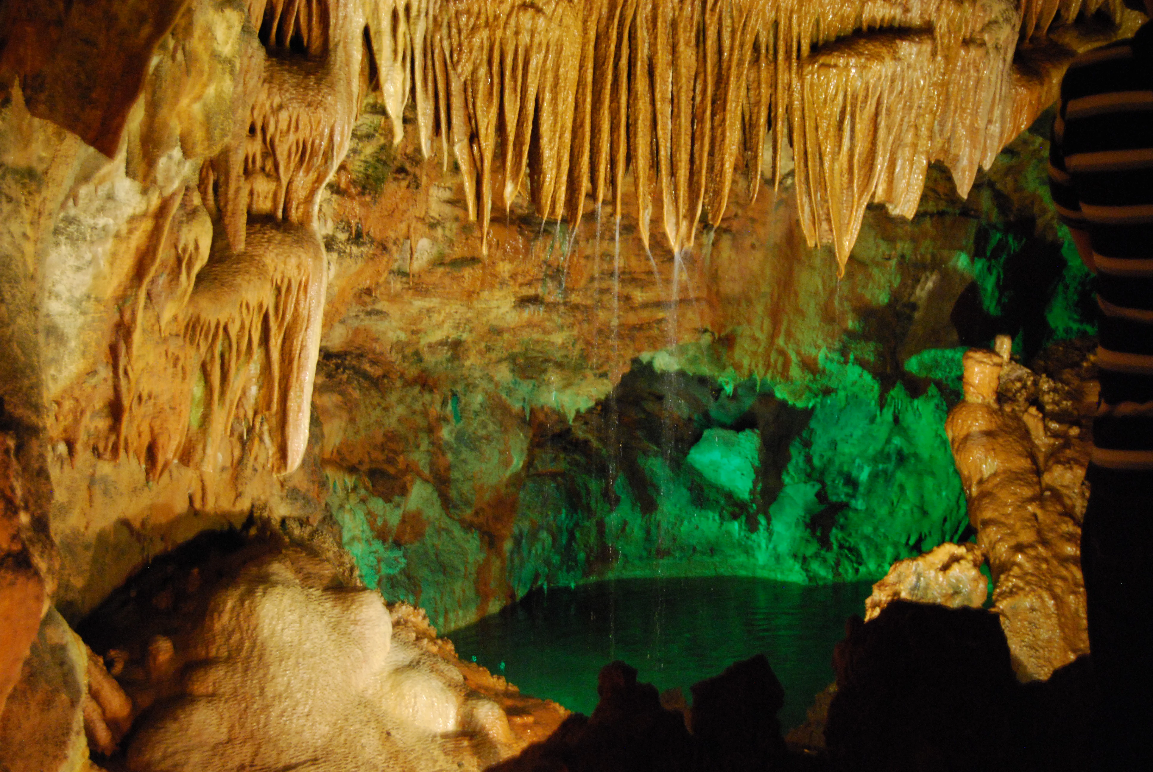 Cave interior with hanging stalactites, rocky formations, and a green-lit water pool
