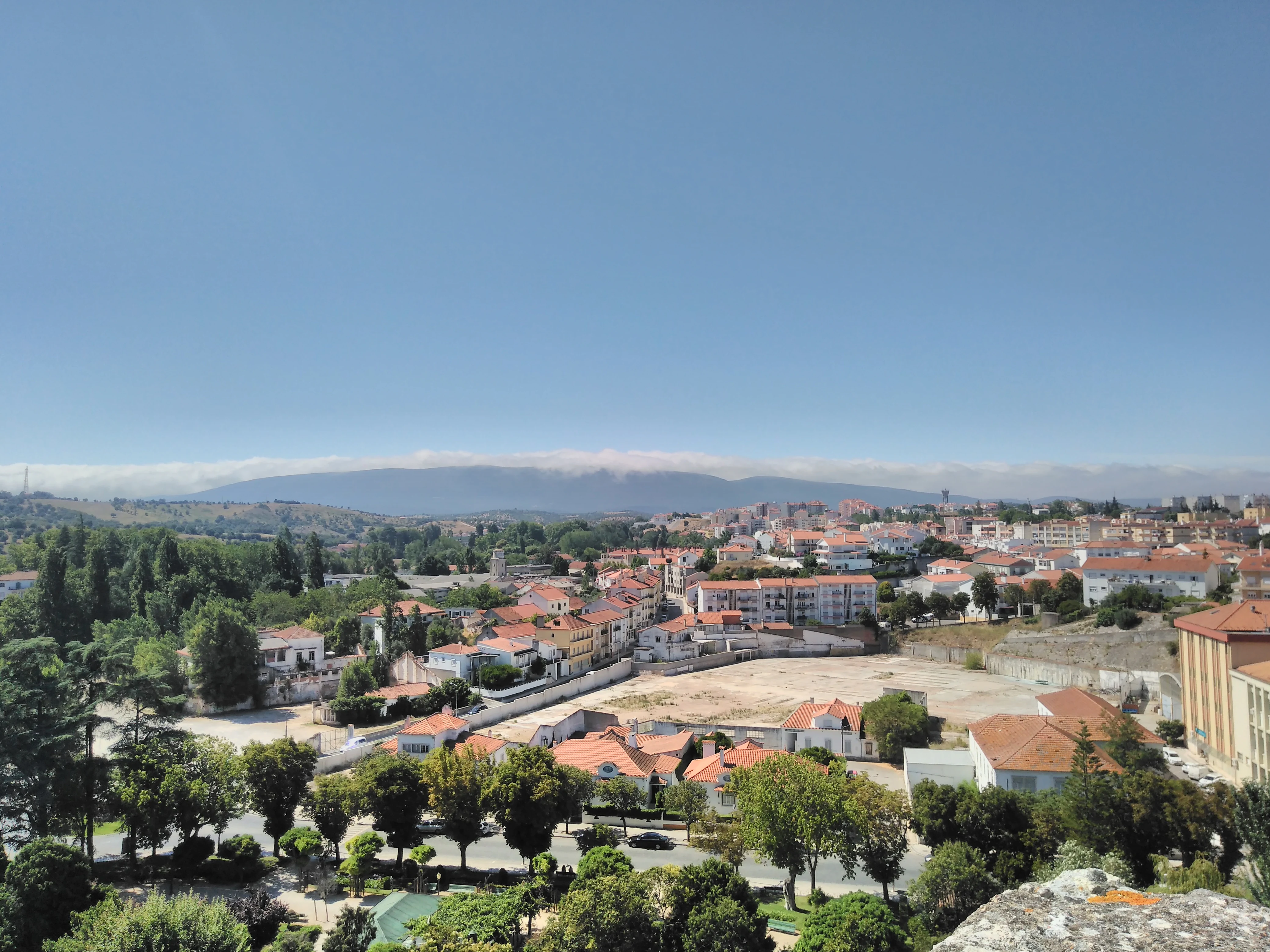 Panoramic view of a town with red-roofed buildings and trees, with the Serras de Aire e Candeeiros mountain range visible in the distance under a clear blue sky