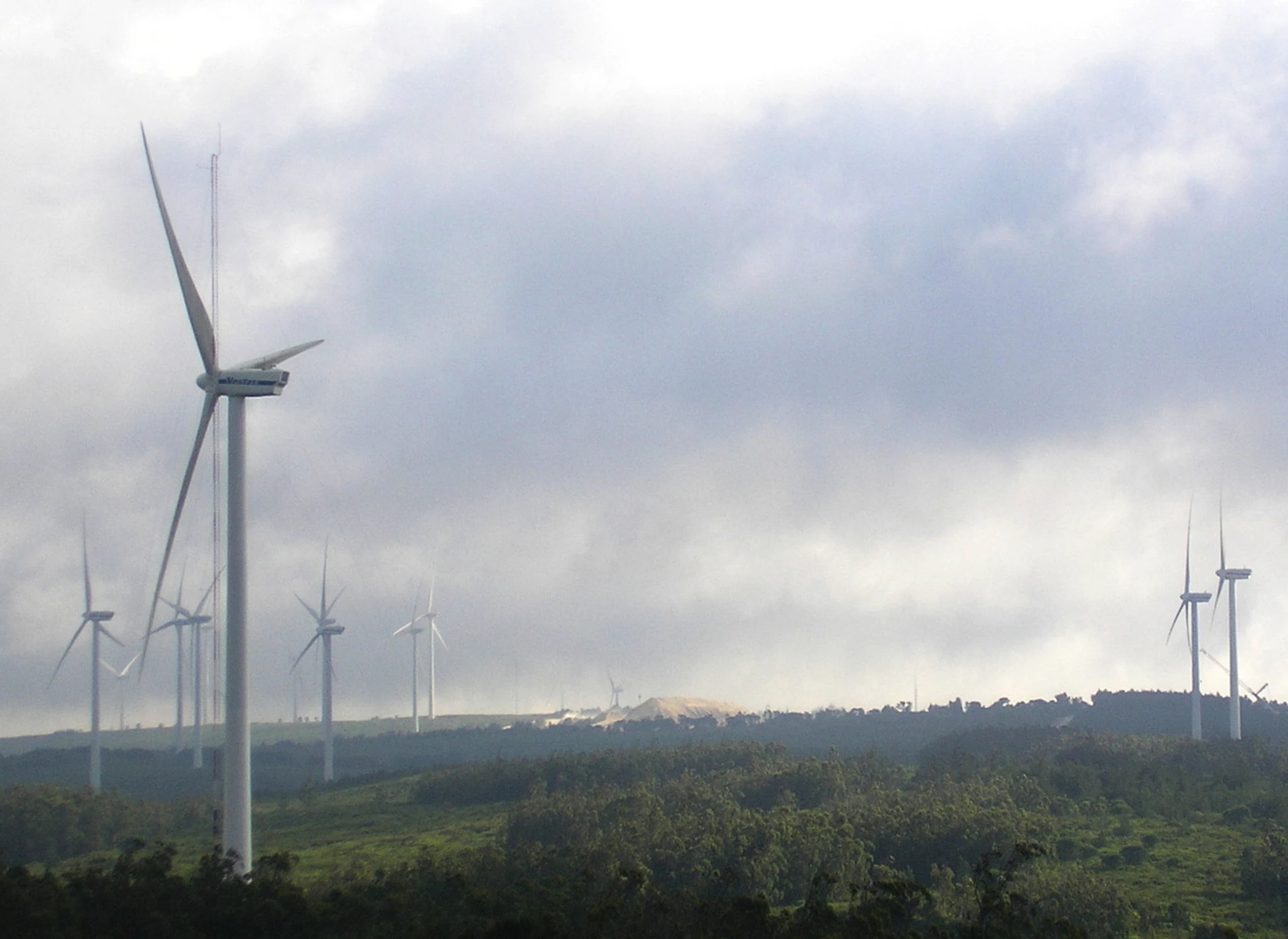 Multiple wind turbines standing on a hillside with dense green vegetation under a cloudy sky
