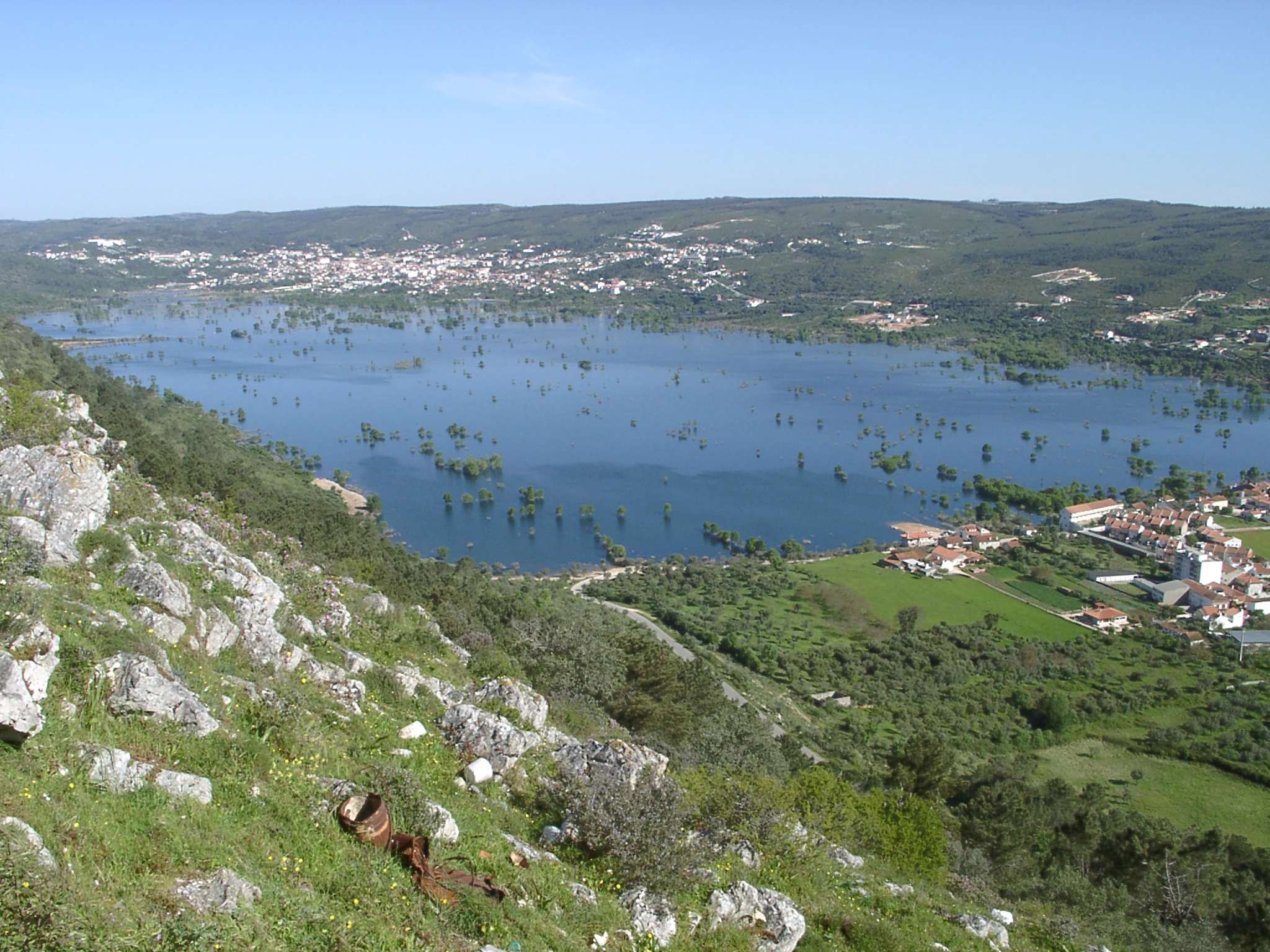 Large body of water surrounded by rocky hills and green vegetation with small settlement visible on right side