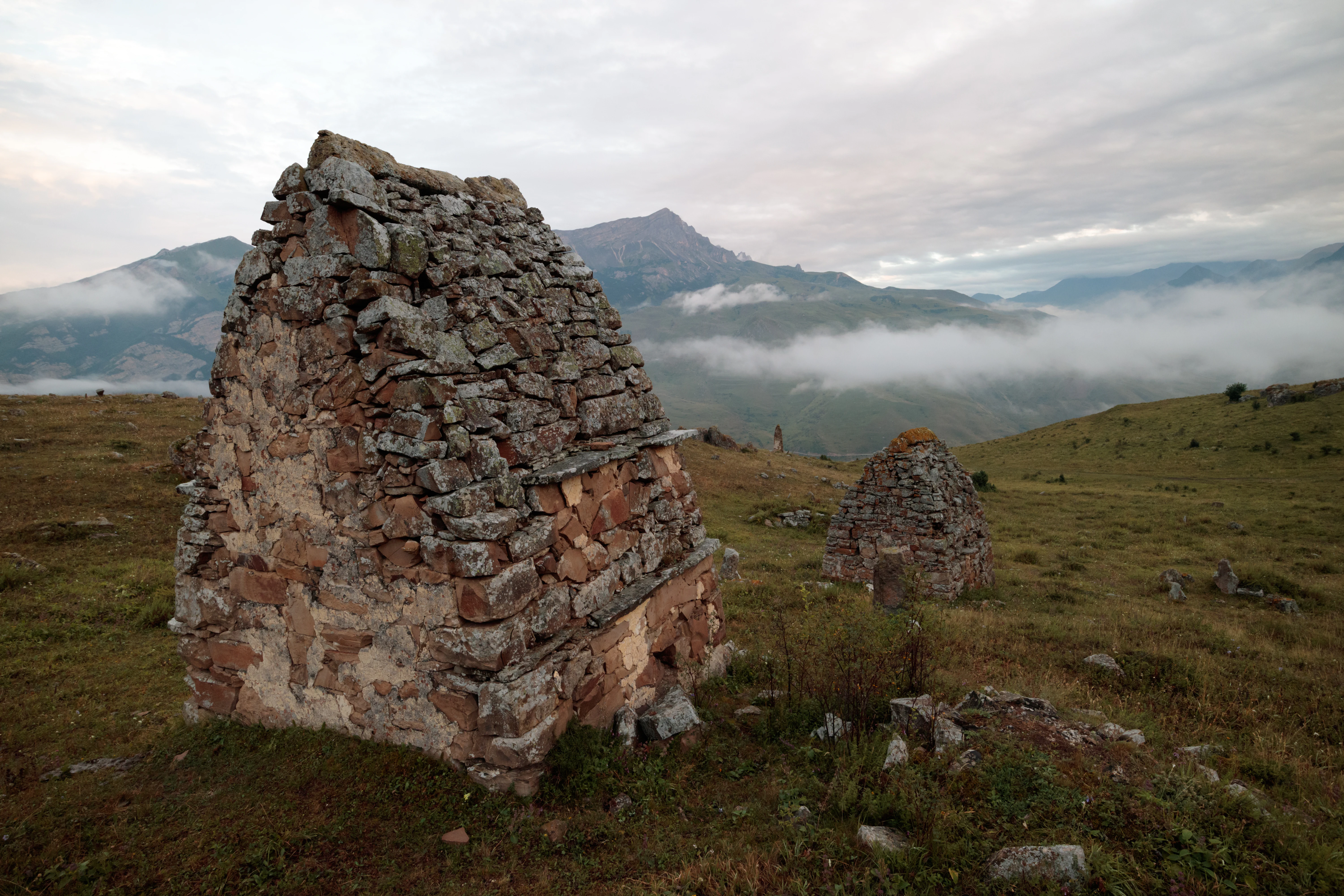 A group of ancient, weathered stone crypts with peaked roofs standing on a grassy, mountainous hillside under a cloudy sky.