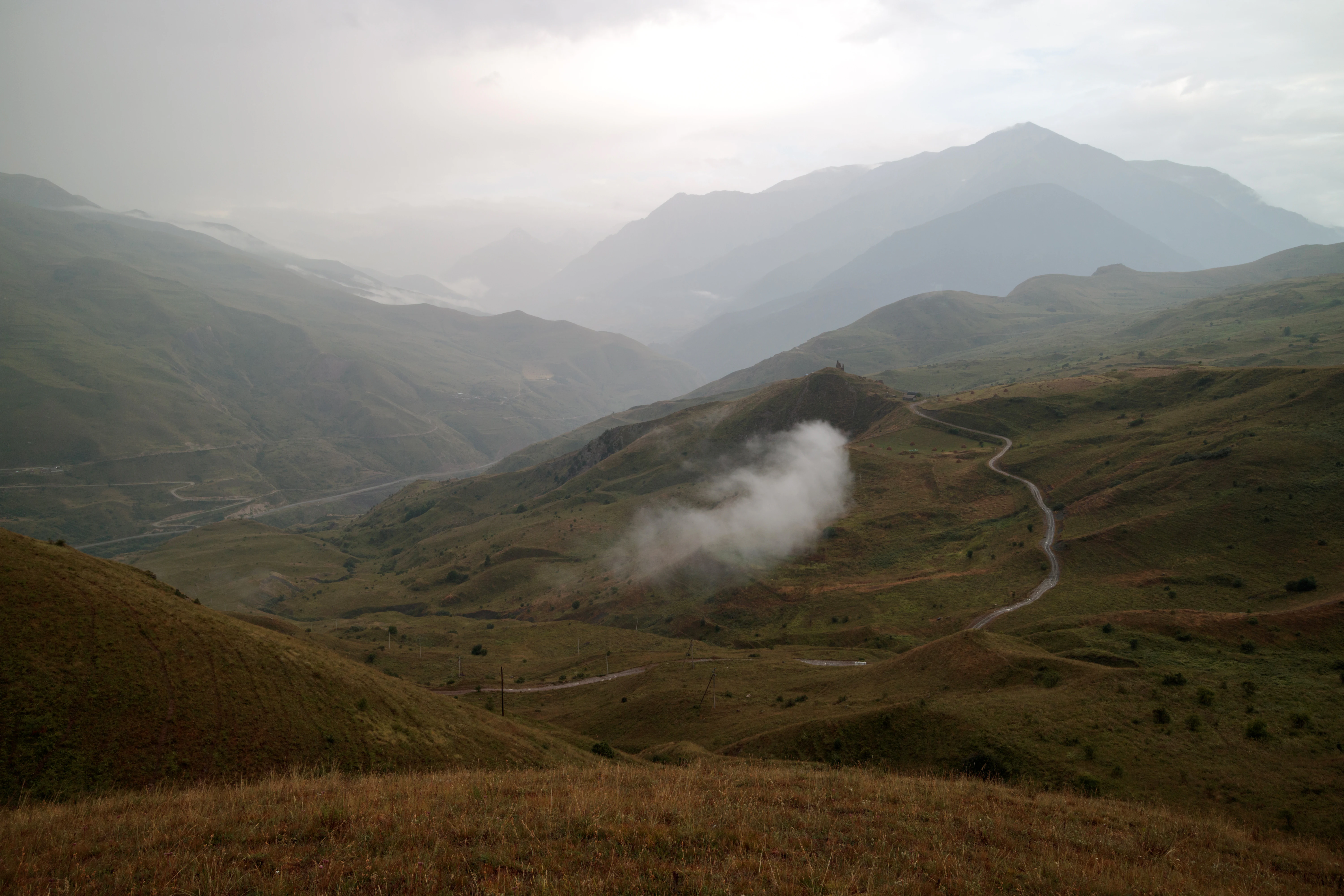 A wide, hazy mountain landscape in the Caucasus with rolling green hills, a winding dirt road in the valley, and low-hanging clouds.