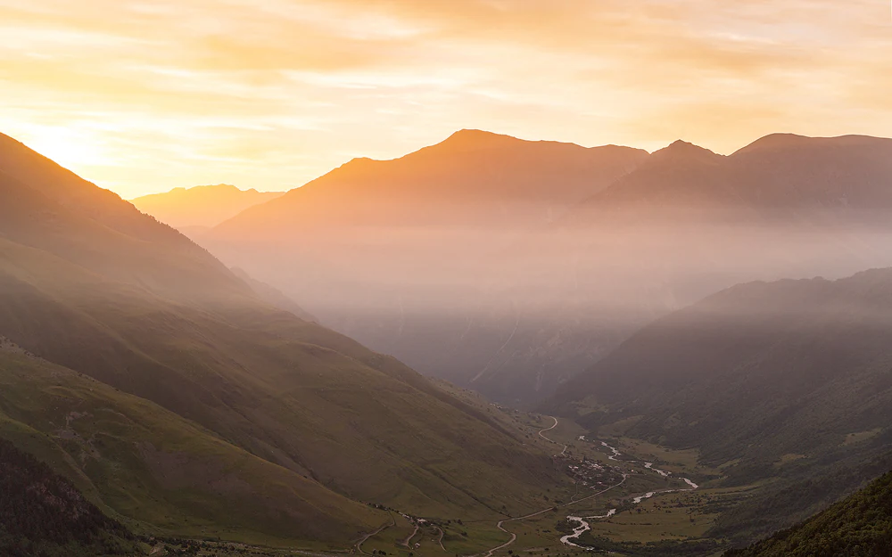 Mountainous landscape at sunrise with valleys, a river, and rolling hills under an orange sky