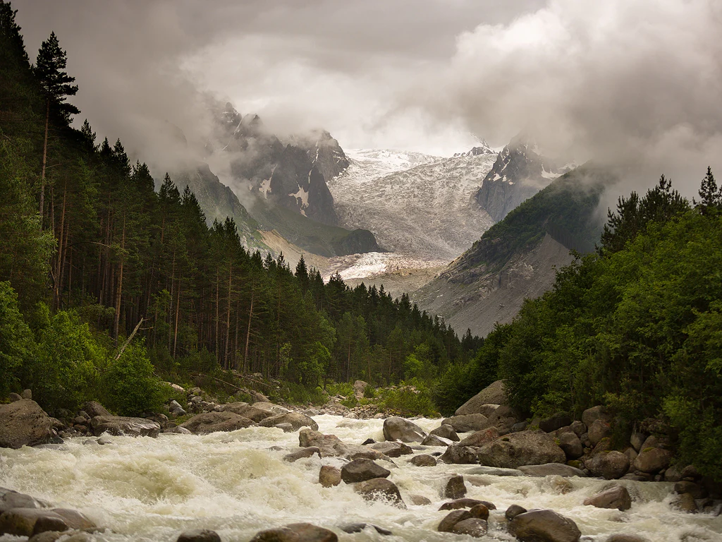 River with rocky rapids flows through a forested gorge, mountains with glacier under cloudy sky