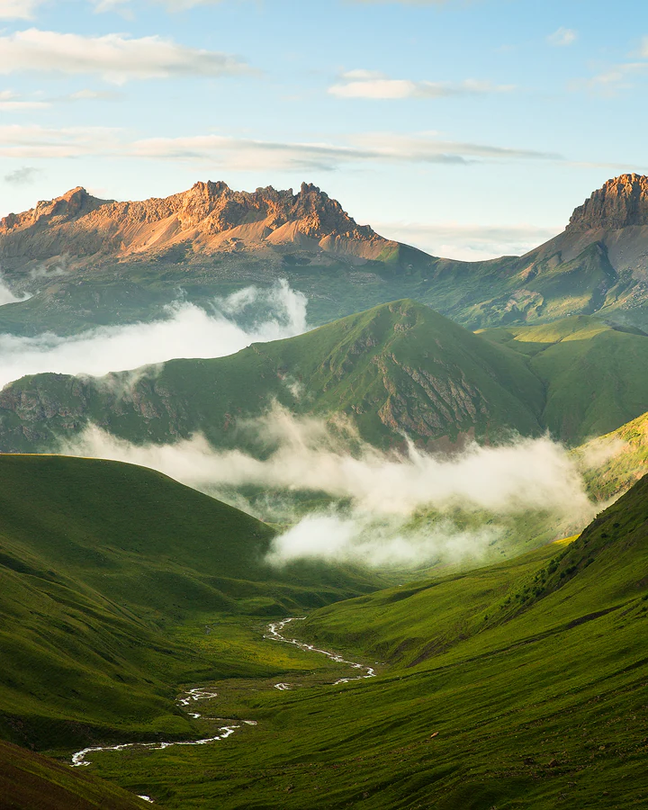 A wide view of a mountainous gorge with green slopes, a winding river, and low-lying clouds between rocky peaks
