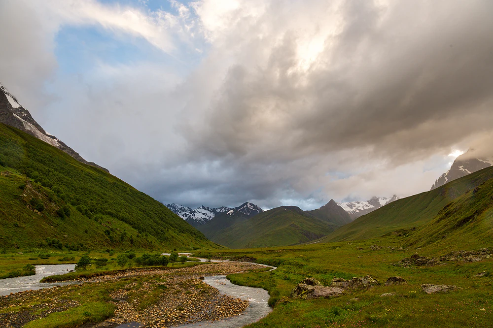 Wide landscape view of a river flowing through a grassy valley with snow-capped mountains in the background under a cloudy sky